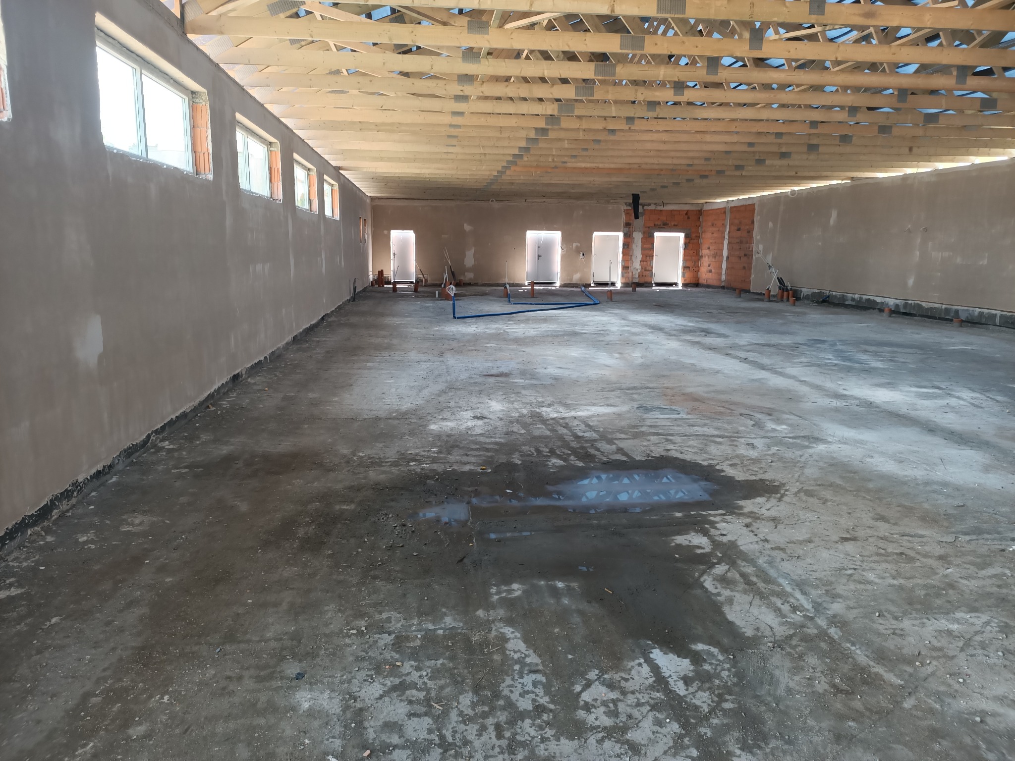 Wnętrze surowego, murowanego budynku w trakcie budowy: widoczne okna, otynkowane ściany, surowa podłoga z plamami wody i drewniana konstrukcja dachu.
