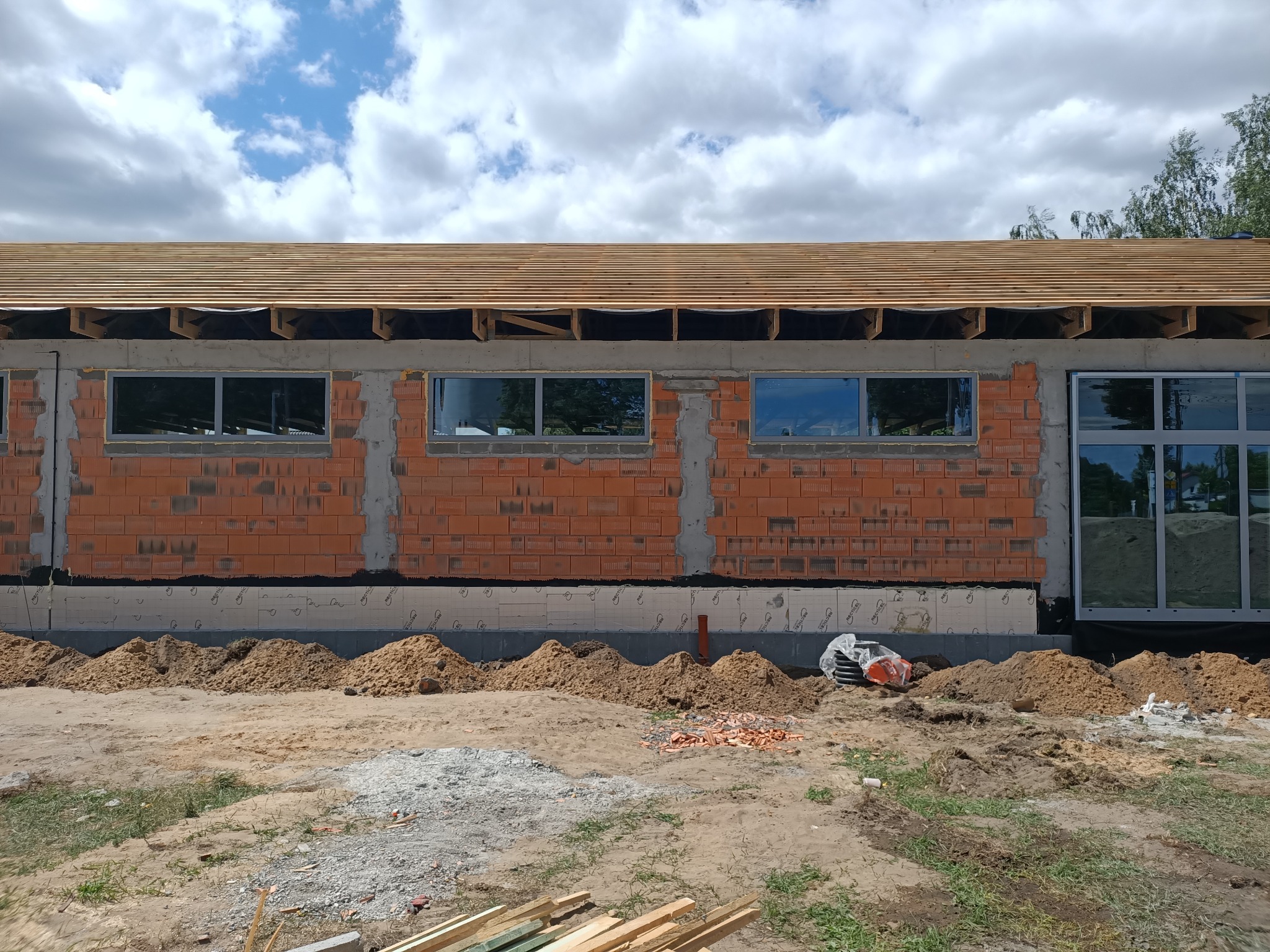 Budowa domu murowanego: ściana z cegły z oknami, widoczny fundament i drewniana konstrukcja dachu w trakcie budowy, piasek i narzędzia na placu budowy.