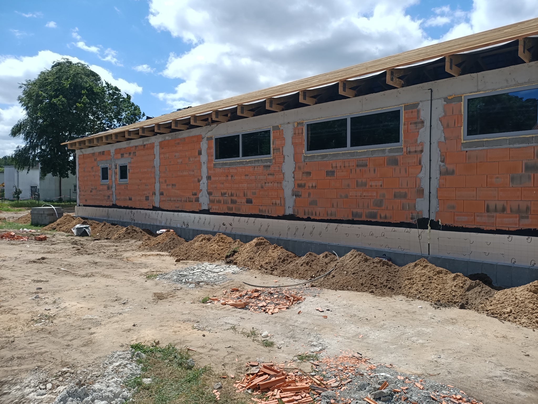 Wznoszony budynek z czerwonej cegły z widocznymi oknami i konstrukcją dachu, otoczony stertami ziemi i gruzu.