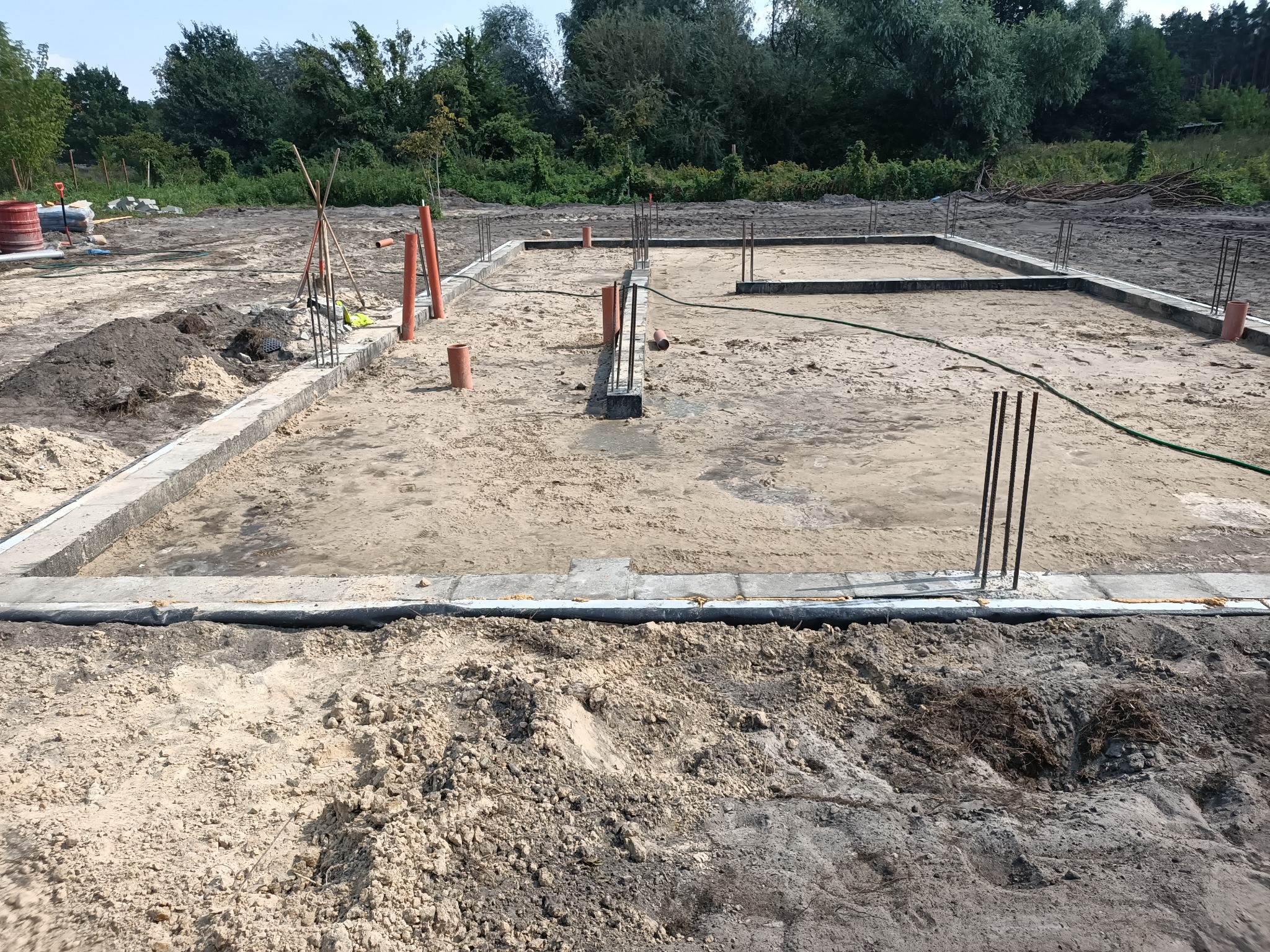 Zalany fundament pod budynek z wystającymi prętami zbrojeniowymi, widoczne rury kanalizacyjne i narzędzia budowlane na piaszczystym terenie.