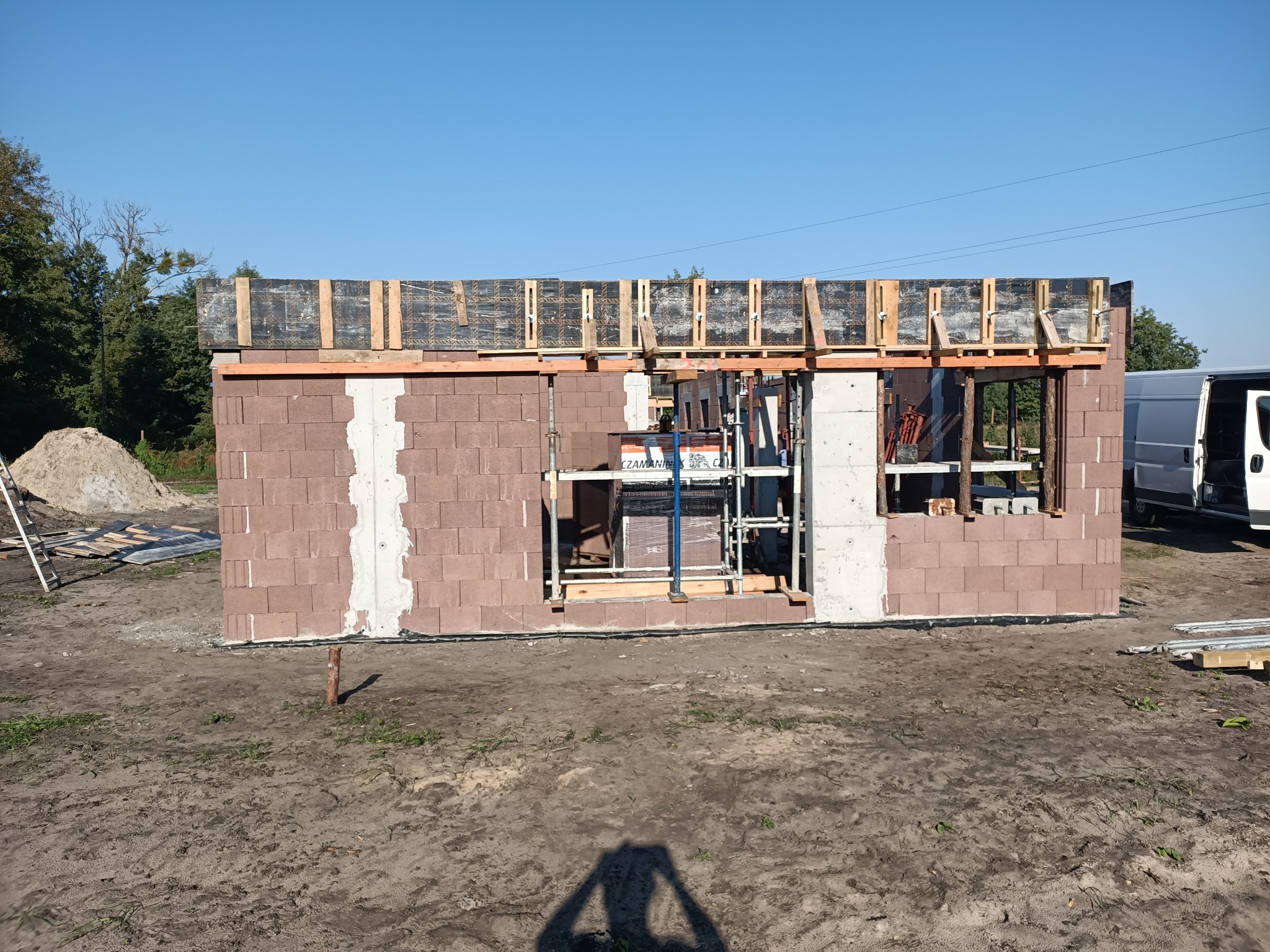 Wznoszone ściany parteru z betonu komórkowego z widocznym zbrojeniem wieńca i szalunkami, na tle błękitnego nieba i zieleni drzew.