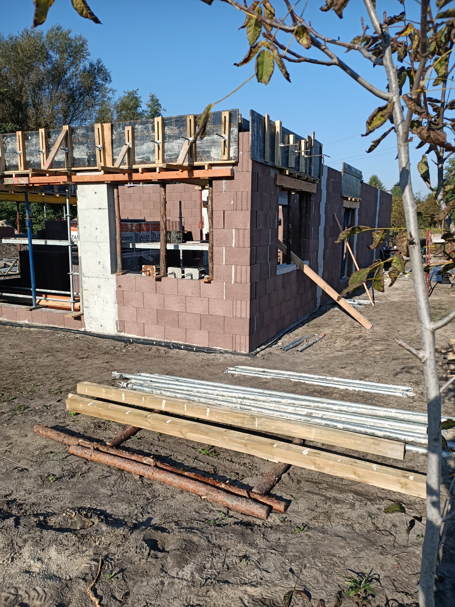 Wznoszenie ścian parterowego budynku z bloczków, widoczne szalunki na betonowe nadproża okienne i strop, w tle rusztowania i błękitne niebo.
