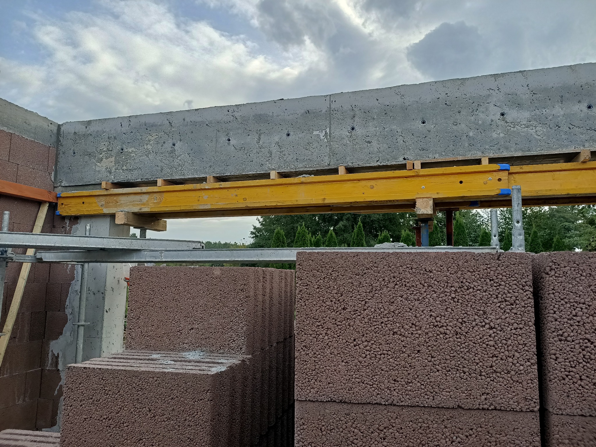 Wznoszenie ściany z czerwonych bloczków betonu komórkowego, widoczne szalunki i podpory stropu betonowego, w tle zieleń drzew i pochmurne niebo.