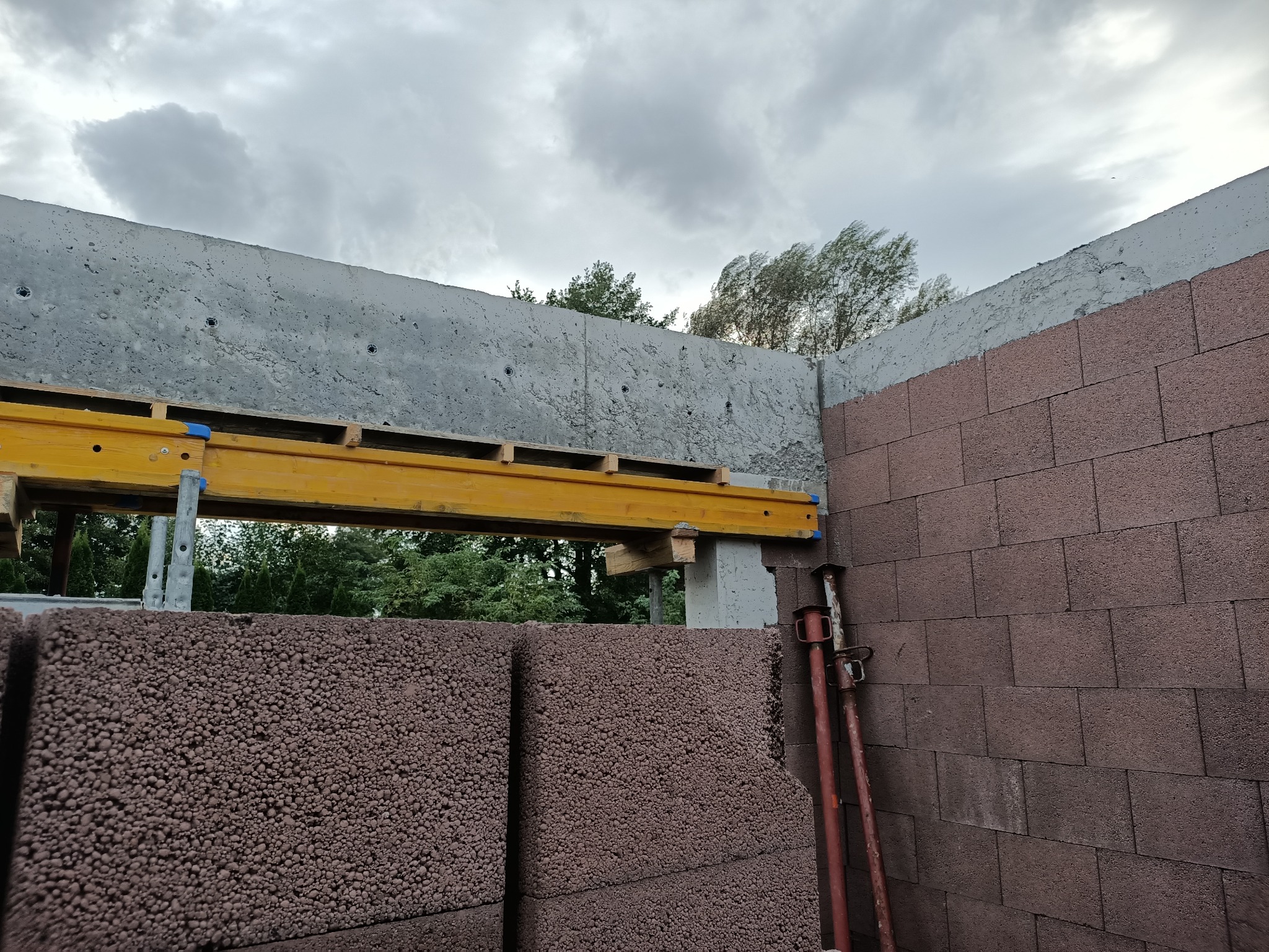 Wznoszenie ściany z brązowych bloczków betonu komórkowego, z widocznym betonowym nadprożem wzmocnionym żółtymi belkami i podporami, w tle zieleń drzew i pochmurne niebo.