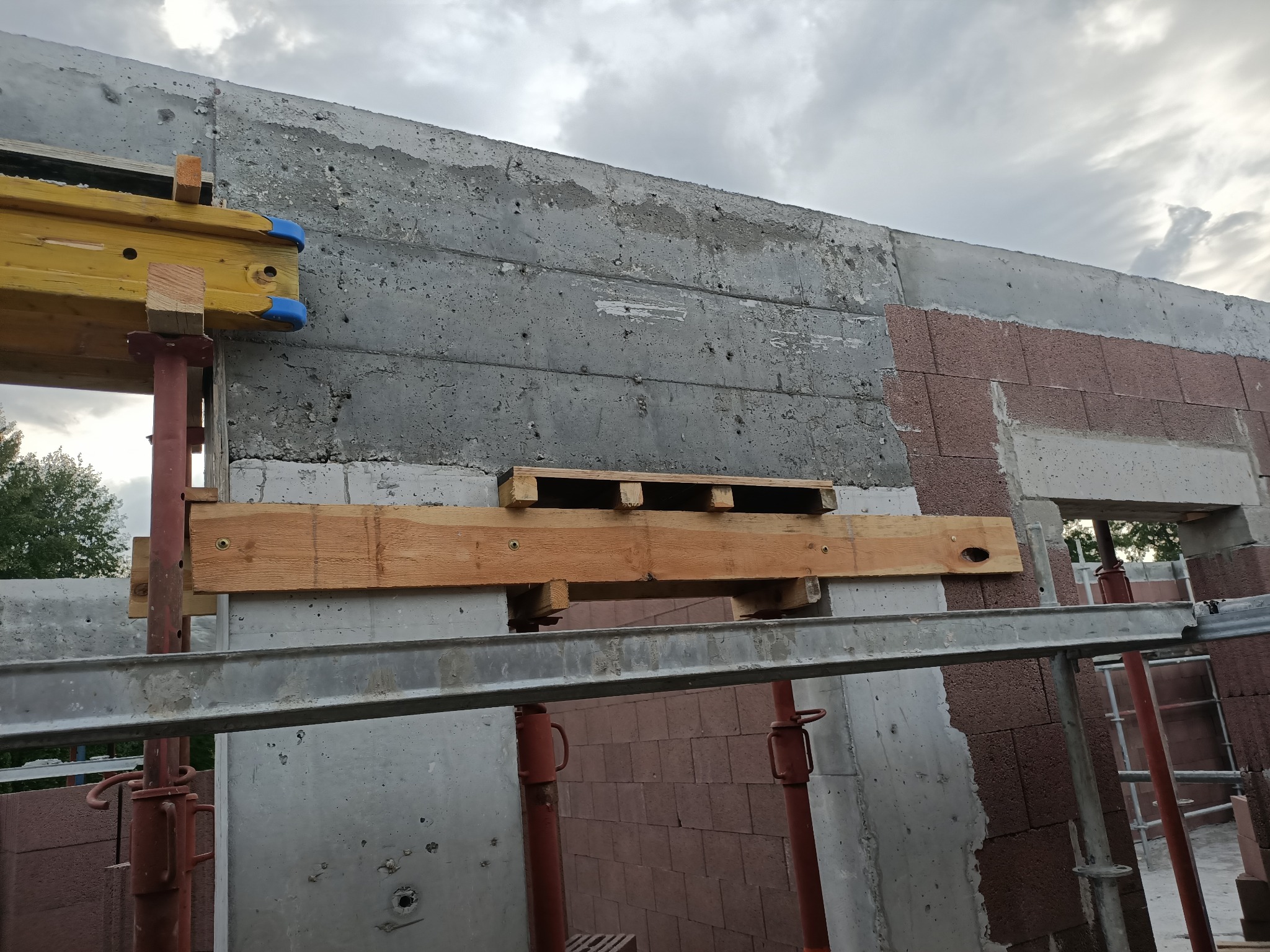 Surowy betonowy mur domu w trakcie budowy, z widocznymi elementami szalunków i podpór, częściowo obłożony czerwoną cegłą, pod pochmurnym niebem.