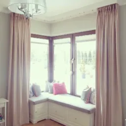 Beżowe zasłony z marszczeniem ołówkowym, dekorujące okno wykuszowe z siedziskiem i poduszkami w odcieniach szarości i różu, oświetlone przez wiszącą lampę z abażurem.