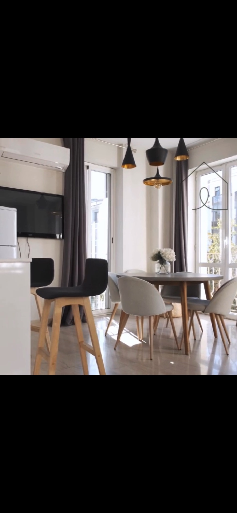 Nowoczesne wnętrze jadalni z szarymi zasłonami, okrągłym stołem, krzesłami w odcieniach szarości i drewnianymi hokerami, oświetlone wiszącymi lampami w kolorze czarnym i złotym.