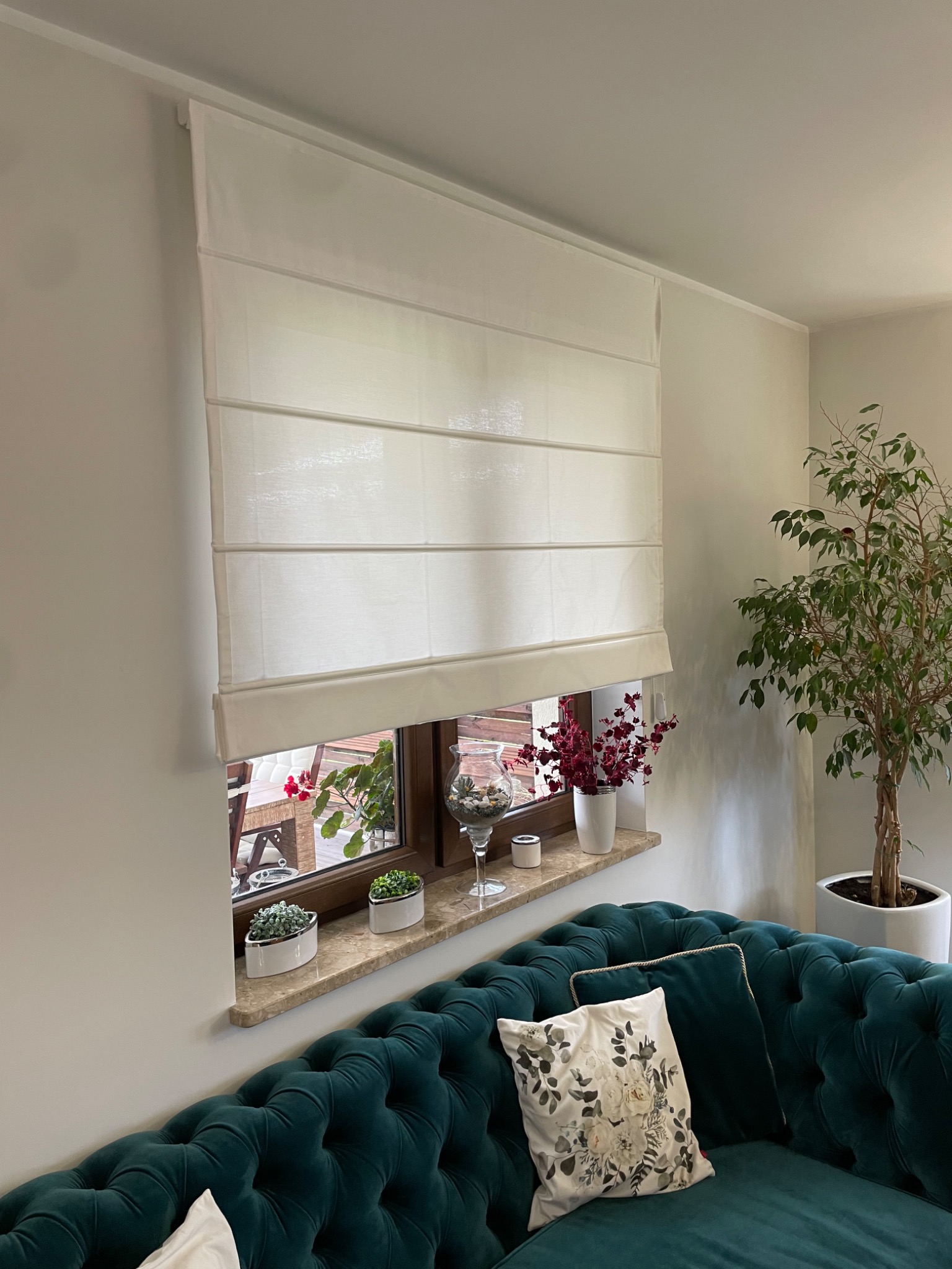 Biała, rzymska roleta na oknie z marmurowym parapetem, na którym stoją dekoracyjne rośliny w doniczkach, widok na zielony ogród z zewnątrz, poniżej turkusowa sofa Chesterfield z ozdobnymi poduszkami.