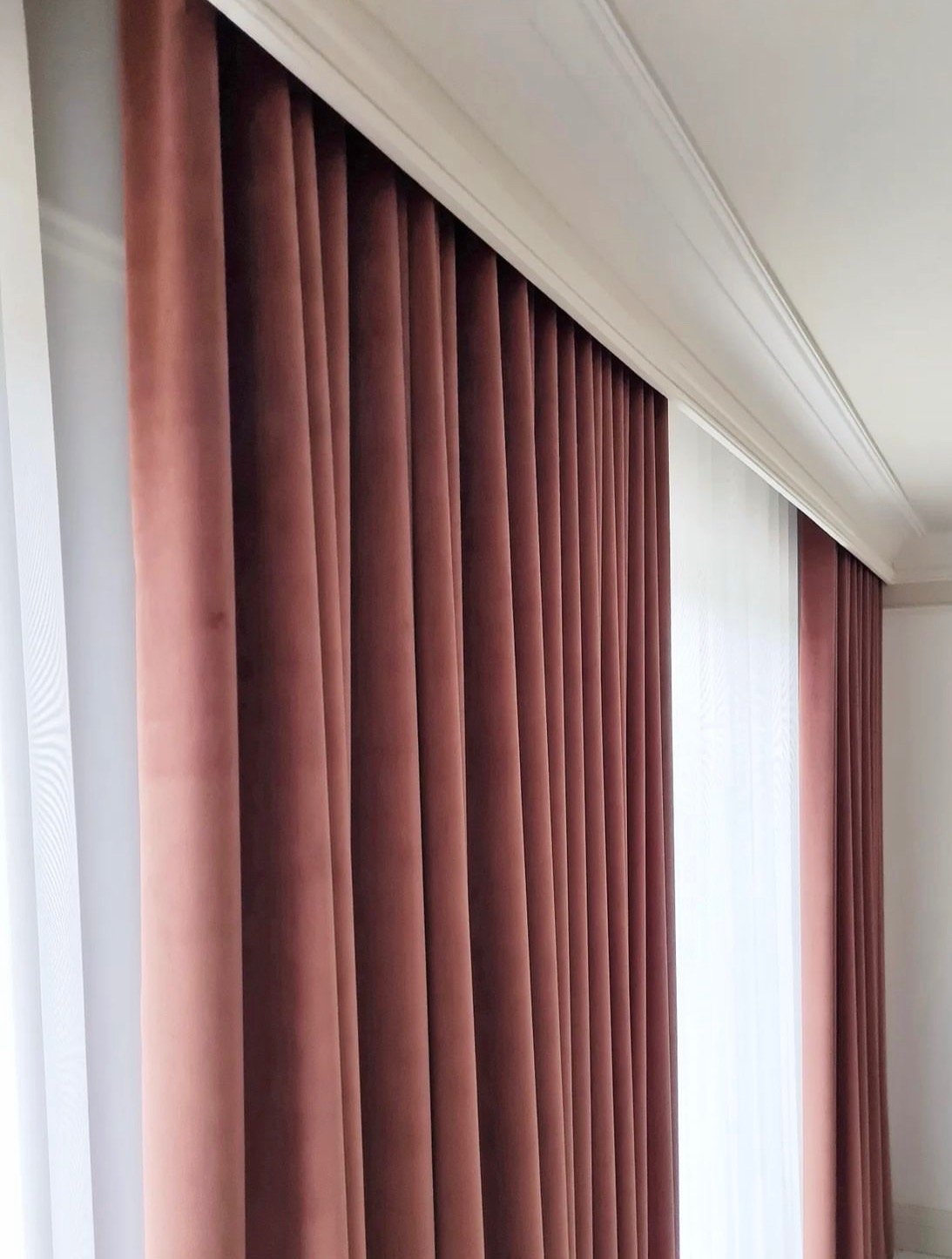 Zasłony z grubego, marszczonego materiału w odcieniu pudrowego różu, wiszące od sufitu do podłogi, z widoczną białą firanką po bokach i dekoracyjną listwą przysufitową.