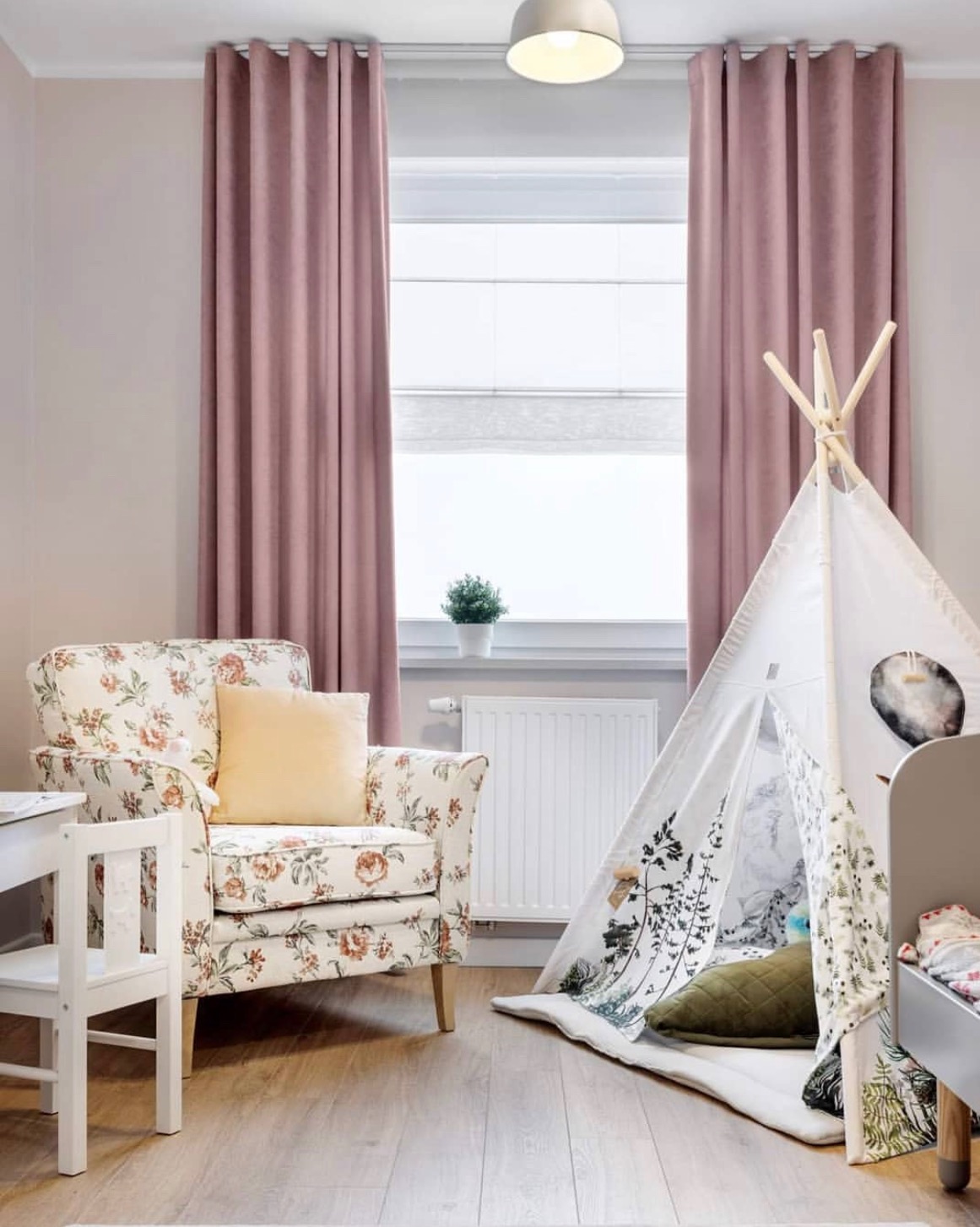 Pokój dziecięcy z zasłonami w odcieniu pudrowego różu, fotel w kwiatowy wzór z żółtą poduszką, tipi z motywem roślinnym i białe meble dziecięce.