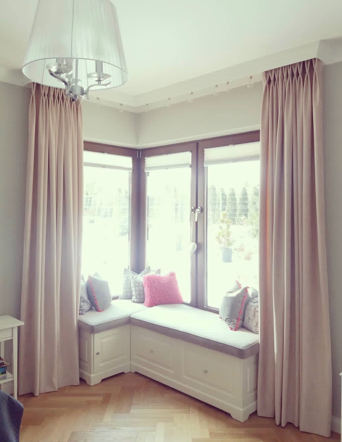 Beżowe zasłony z marszczeniem ołówkowym, dekorujące okno wykuszowe z siedziskiem i poduszkami w odcieniach szarości i różu, oświetlone przez wiszącą lampę z abażurem.