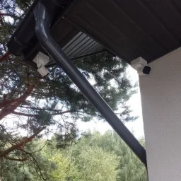 Dwie białe kamery monitoringu zamontowane pod okapem budynku, widoczne rynna spustowa i fragment drzewa iglastego na tle zieleni.