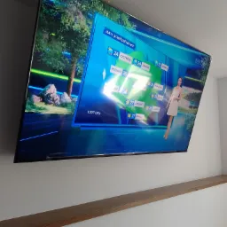 Telewizor 85 cali zamontowany na ścianie nad drewnianą półką, wyświetlający prognozę pogody TVP.