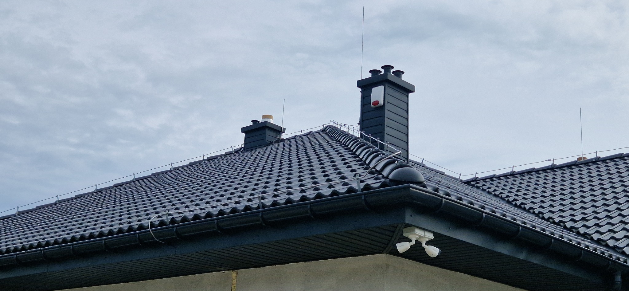 Czarny dach z dachówki ceramicznej, dwa kominy (jeden z czerwoną obudową alarmową), instalacja odgromowa i dwie białe kamery monitoringu pod okapem.