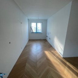 Malowanie mieszkań i lokali firmowych - Oleksandr Shulyar - Pusty pokój w Warszawie z parkietem ułożonym we wzór jodełki, świeżo wykończone ściany, przygotowane punkty elektryczne, widoczne promienie słoneczne wpadające przez okno.