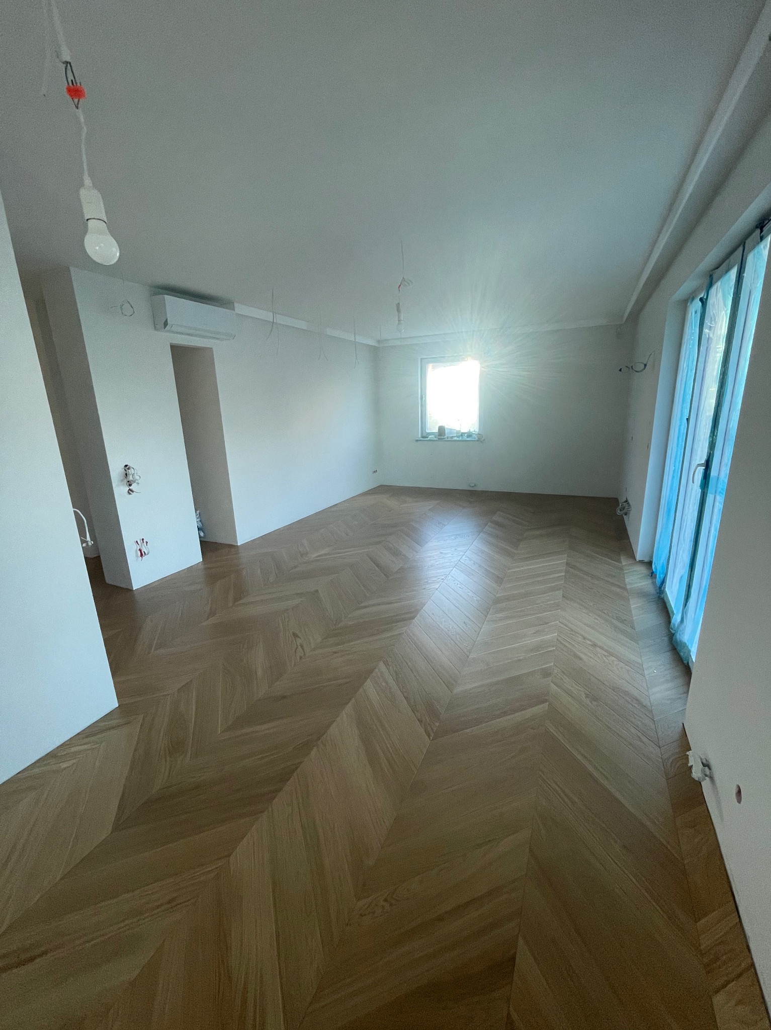 Puste pomieszczenie po remoncie z parkietem ułożonym we wzór jodełki; okno z widocznym światłem słonecznym, białe ściany z widocznymi przewodami elektrycznymi, klimatyzator na ścianie, drzwi...