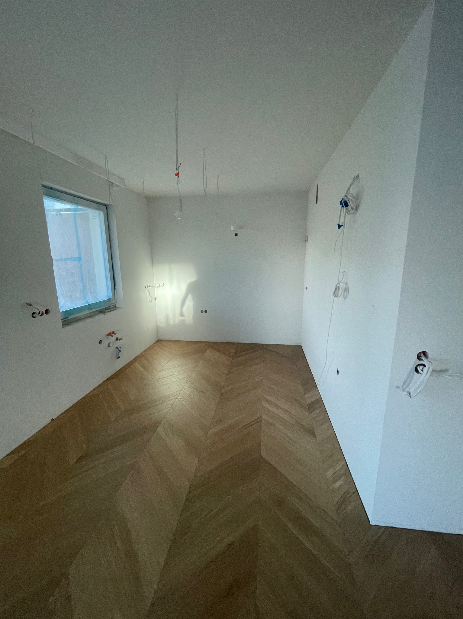 Pusty pokój z parkietem ułożonym we wzór jodełki, surowe ściany z wystającymi przewodami elektrycznymi, okno zabezpieczone folią malarską.