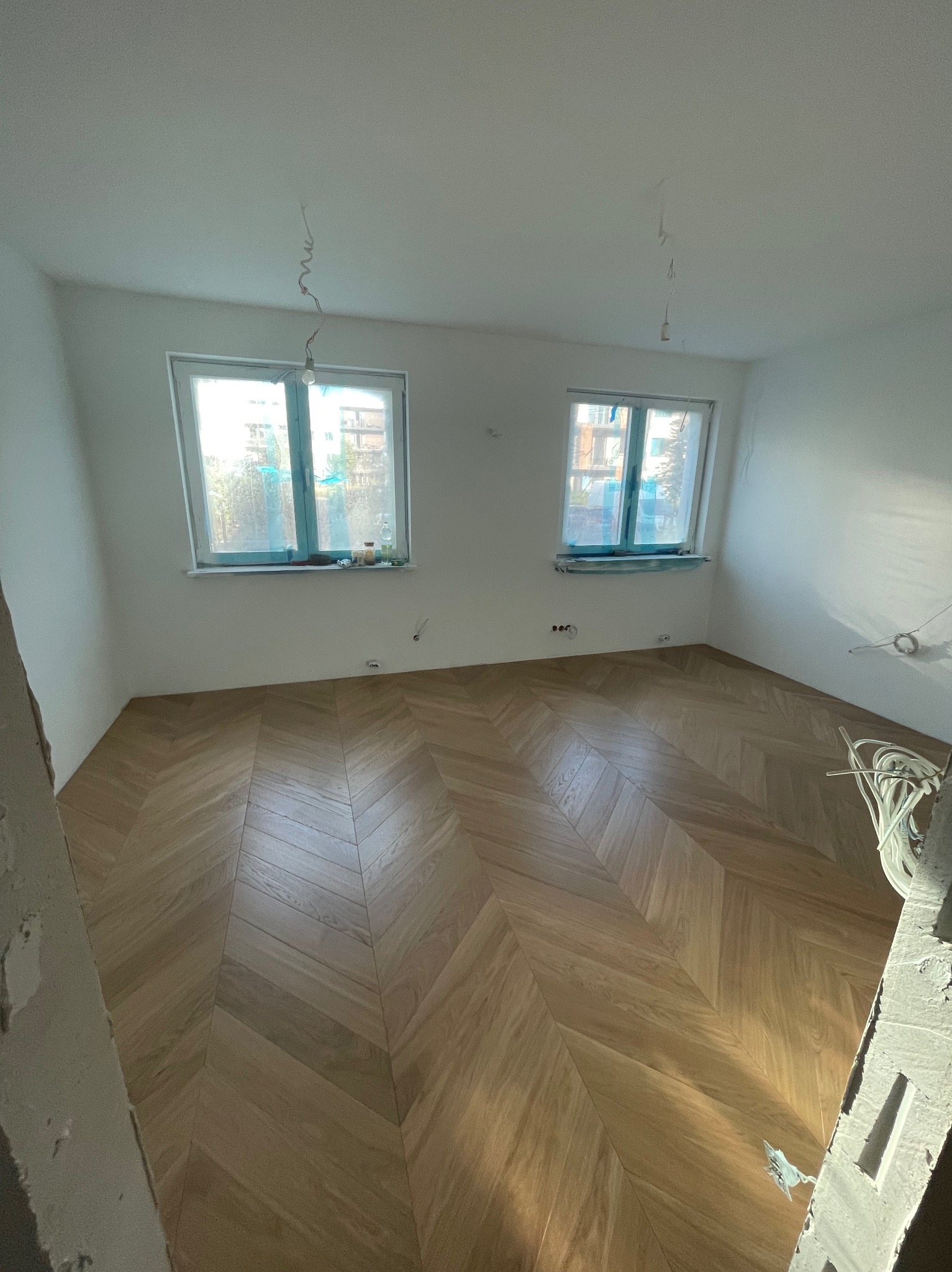 Pokój w trakcie remontu z ułożoną parkietową podłogą w jodełkę, widoczne dwa okna zabezpieczone folią malarską i wystające kable elektryczne.
