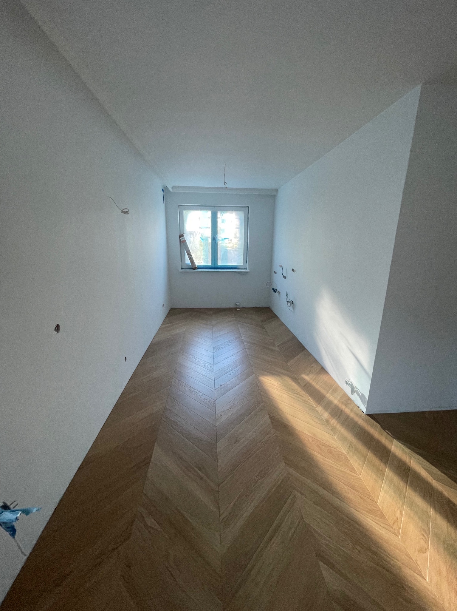 Pusty pokój w Warszawie z parkietem ułożonym we wzór jodełki, świeżo wykończone ściany, przygotowane punkty elektryczne, widoczne promienie słoneczne wpadające przez okno.