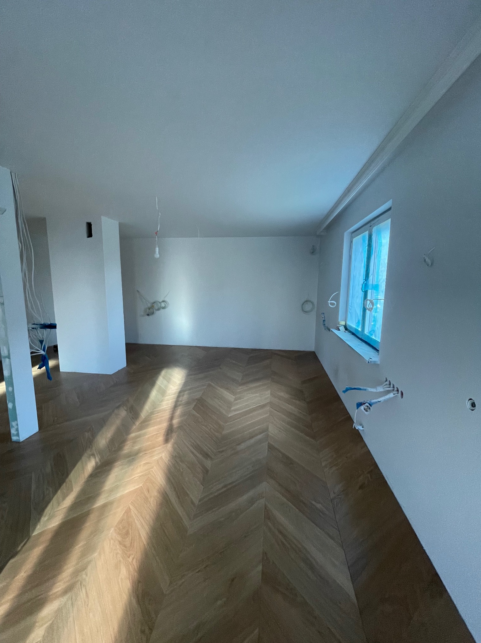 Wnętrze w trakcie wykańczania: świeżo położony parkiet w jodełkę, białe ściany z wystającymi przewodami elektrycznymi, okno zabezpieczone folią malarską.