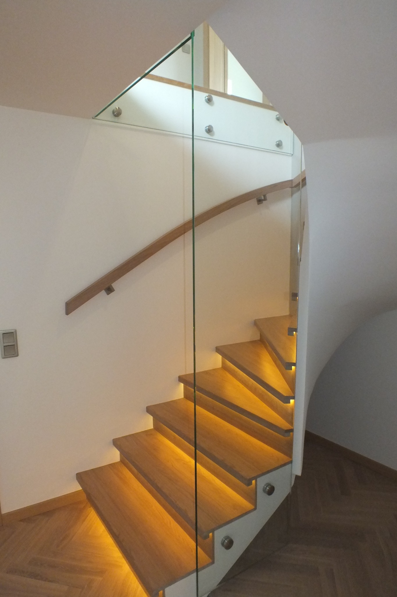 Nowoczesne schody drewniane z podświetleniem LED stopni i szklaną balustradą mocowaną punktowo, widok z dołu na zakręcające schody.