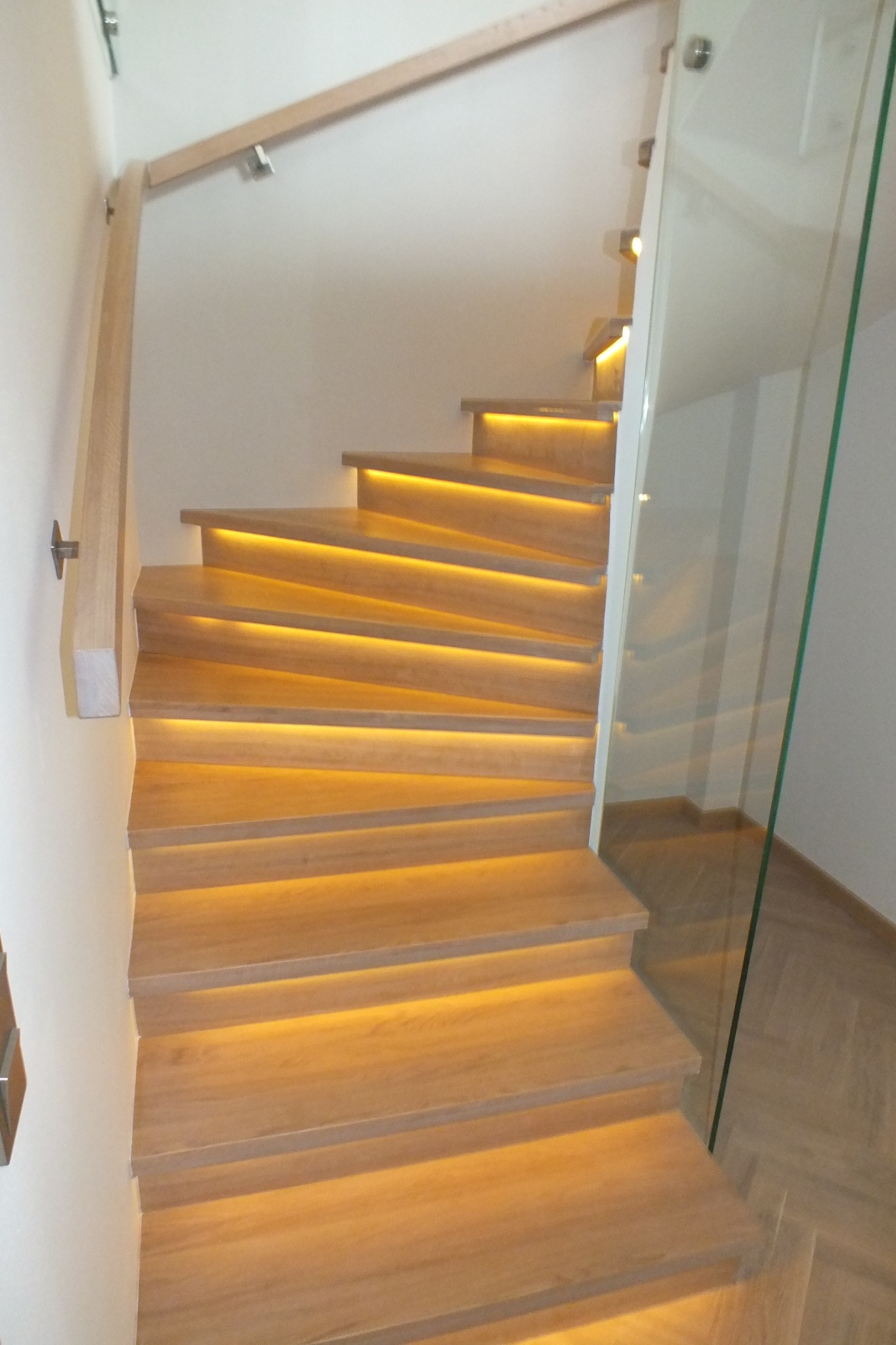 Drewniane schody kręcone z podświetleniem LED pod każdym stopniem, balustrada ze szkła hartowanego, widok z dołu.