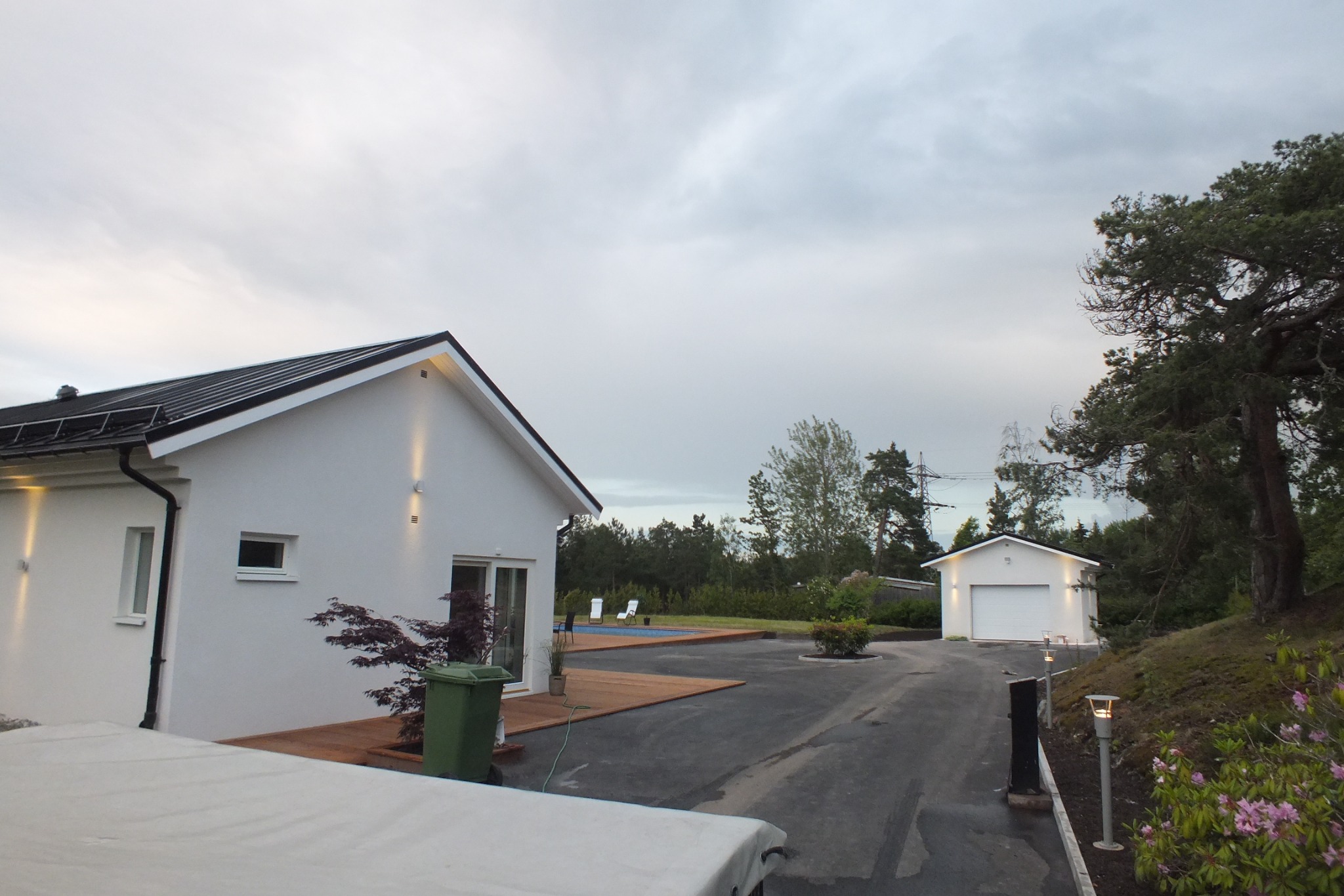 Nowoczesny, parterowy dom z białymi ścianami i czarnym dachem oraz garaż wolnostojący, widoczne z zewnątrz oświetlenie elewacyjne, podjazd i ogród z basenem w tle.