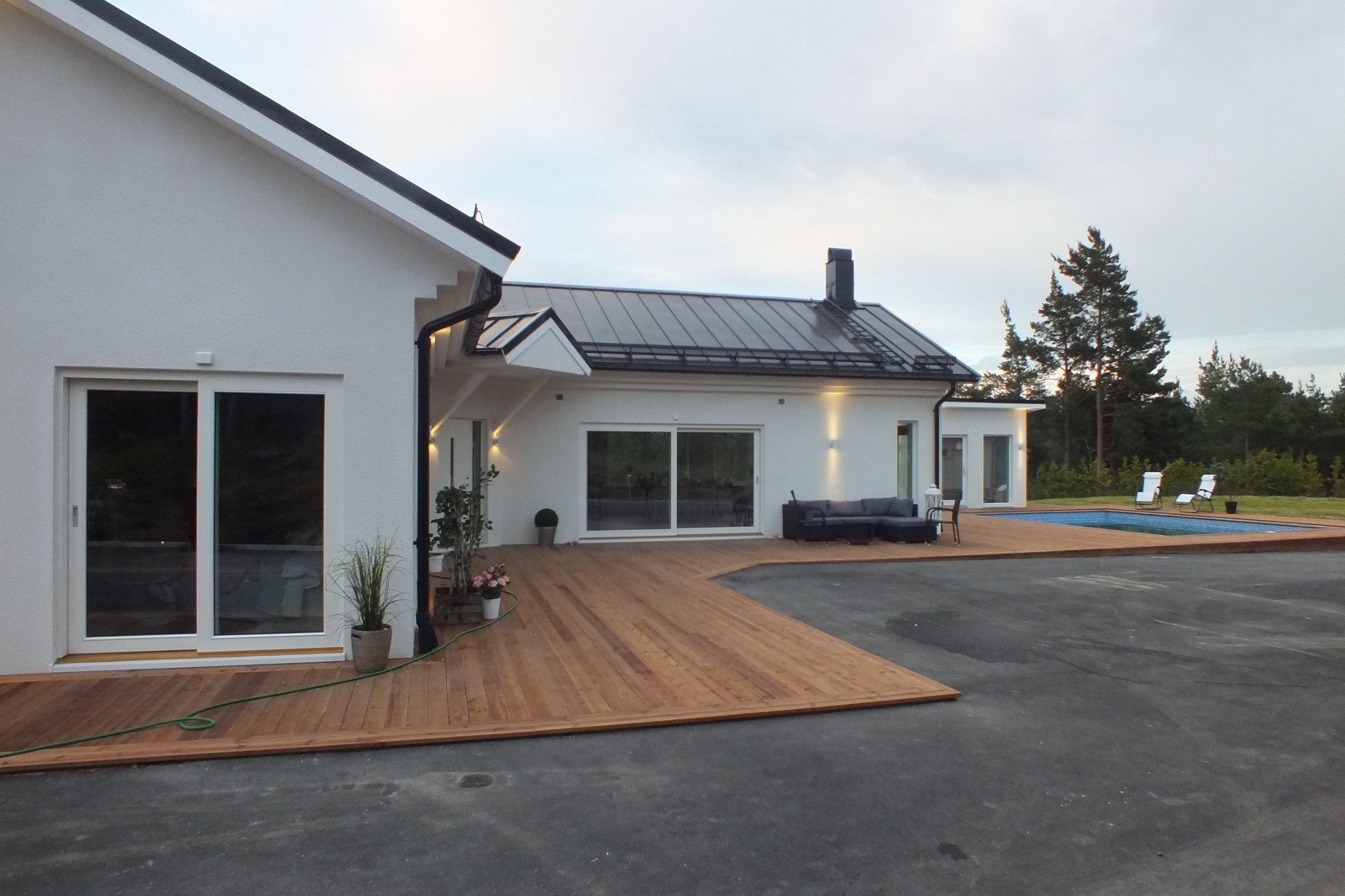 Nowoczesny, parterowy dom z białymi ścianami, ciemnym dachem i dużymi przesuwnymi drzwiami tarasowymi, połączony drewnianym tarasem z basenem; widoczne meble ogrodowe i rośliny w donicach.