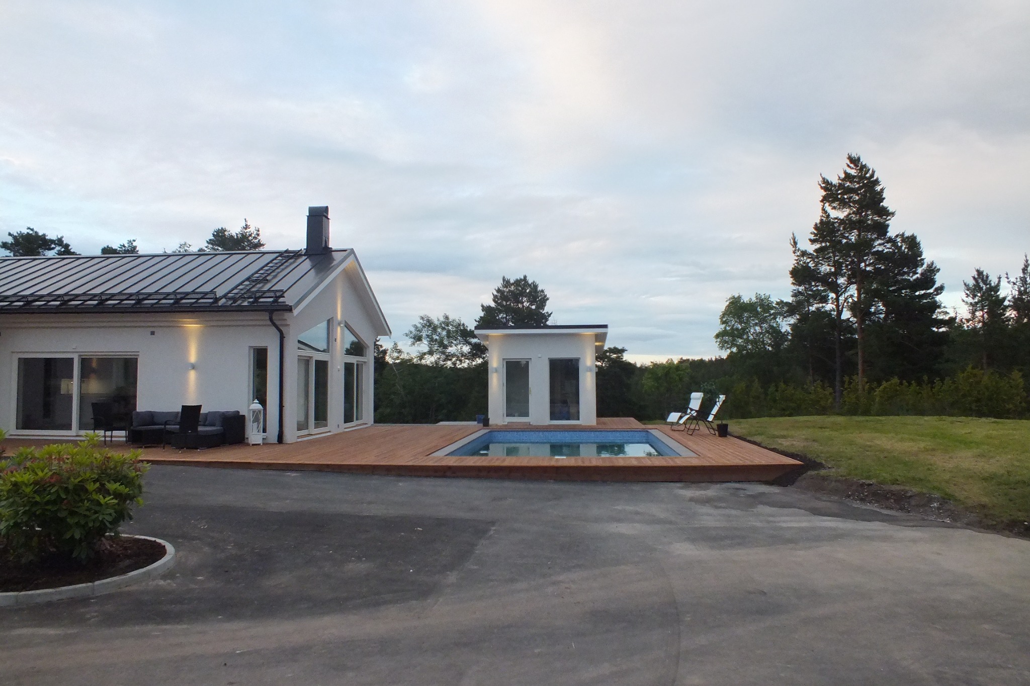 Nowoczesny dom z basenem zewnętrznym i drewnianym tarasem, biała elewacja, metalowy dach, widok z podjazdu.