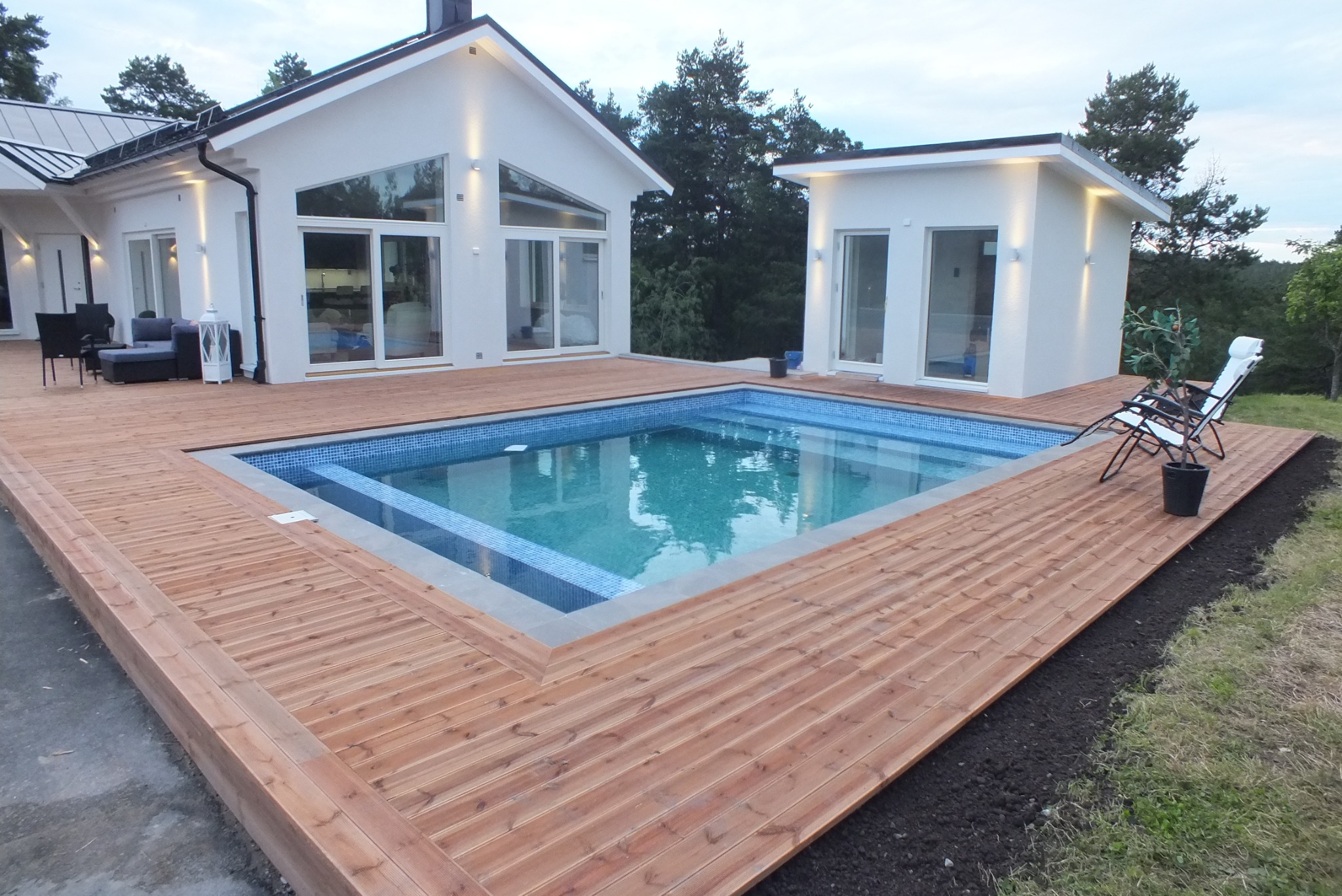 Nowoczesny dom z basenem otoczonym drewnianym tarasem; widoczne duże przesuwne okna, minimalistyczne oświetlenie zewnętrzne i leżaki przy basenie.