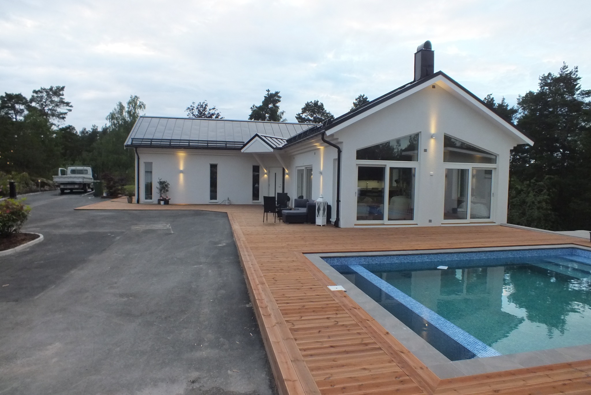 Nowoczesny dom jednorodzinny z białymi ścianami, dużymi oknami i drewnianym tarasem otaczającym basen, z widocznym podjazdem i samochodem dostawczym w tle.