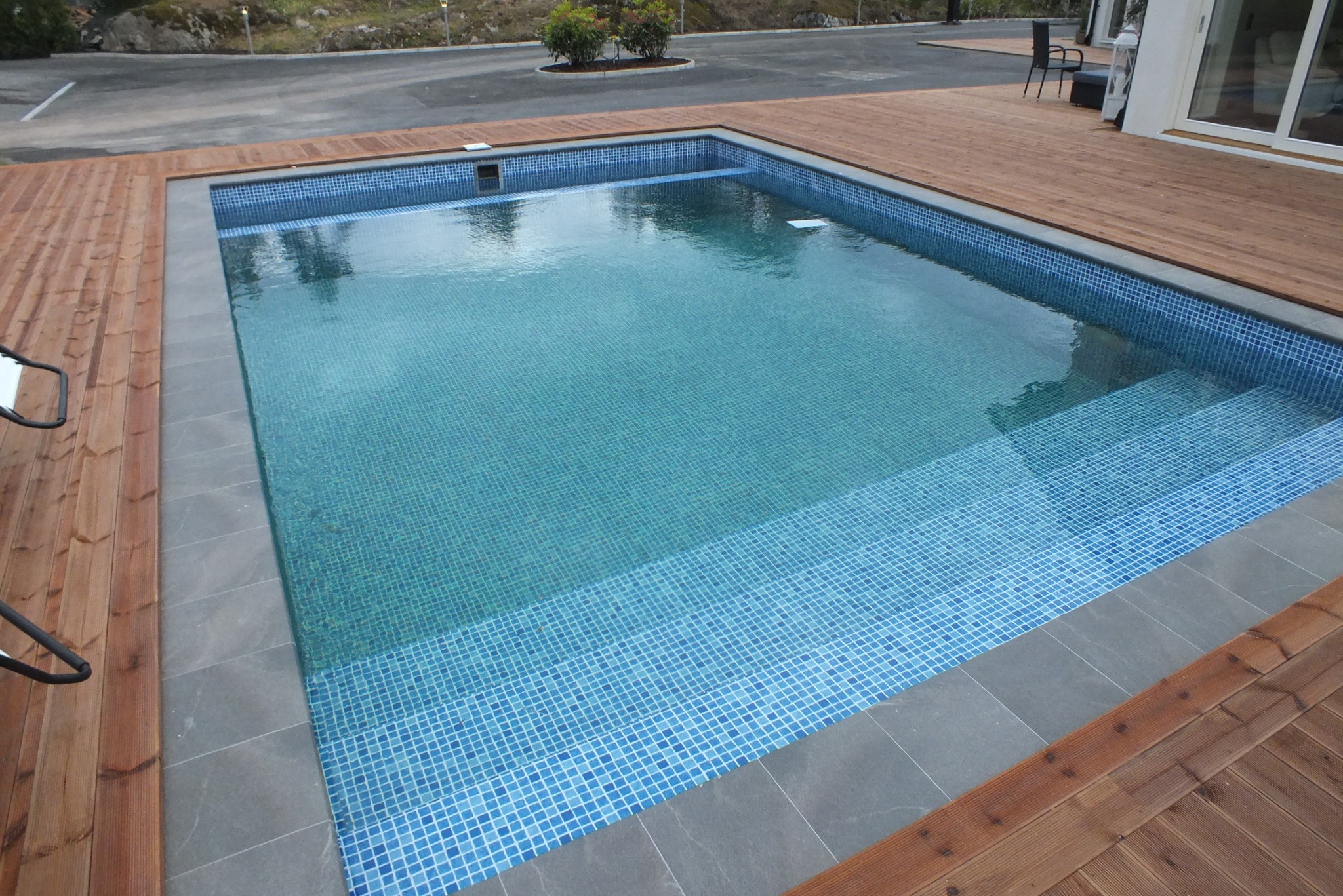 Prostokątny basen z niebieską mozaiką, otoczony szarą płytką i drewnianym tarasem, widok z góry w słoneczny dzień.