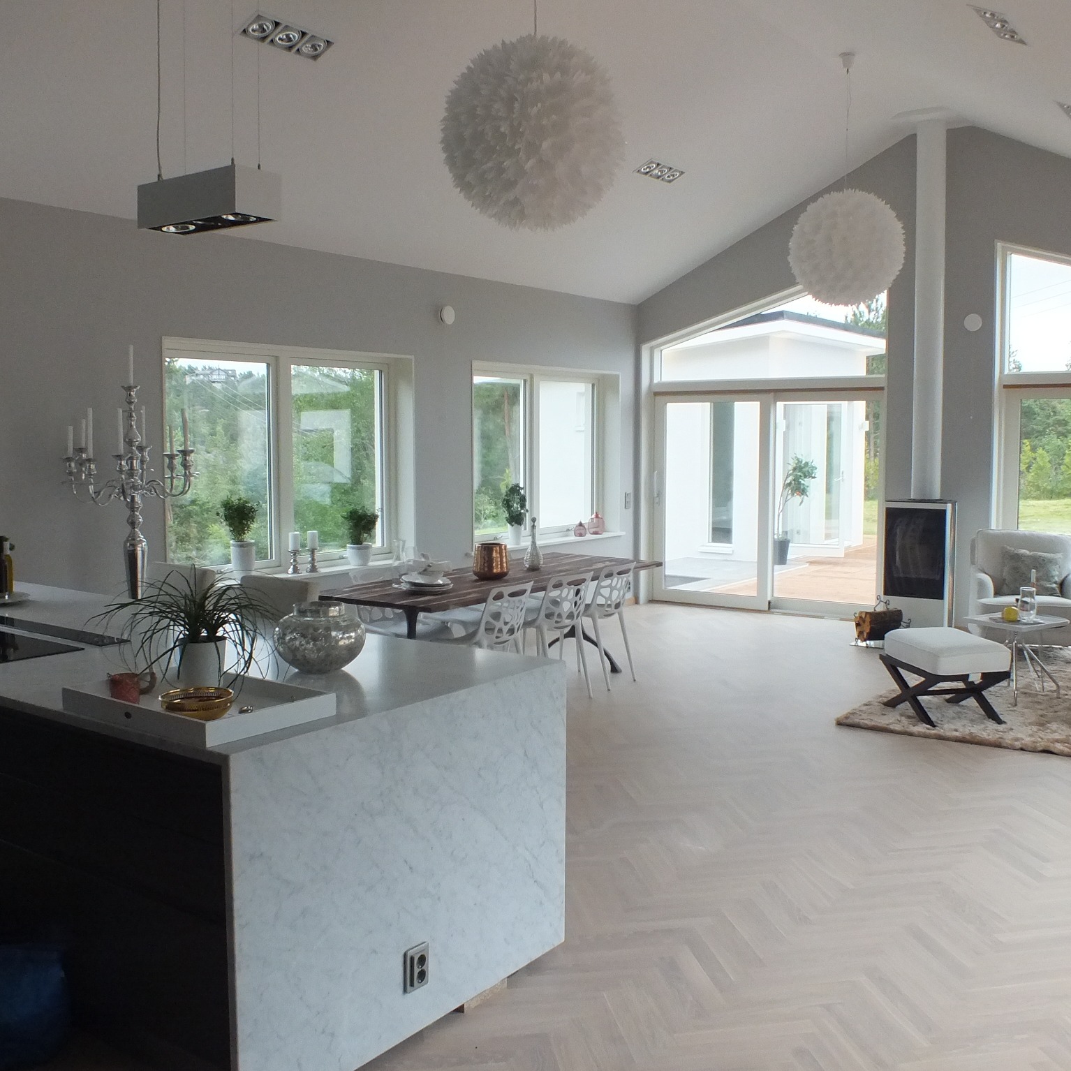 Jasne, przestronne wnętrze domu z dużymi oknami, drewnianą podłogą ułożoną w jodełkę, wyspą kuchenną z marmurowym blatem, stołem jadalnym z białymi krzesłami oraz wiszącymi lampami w kształcie kul.