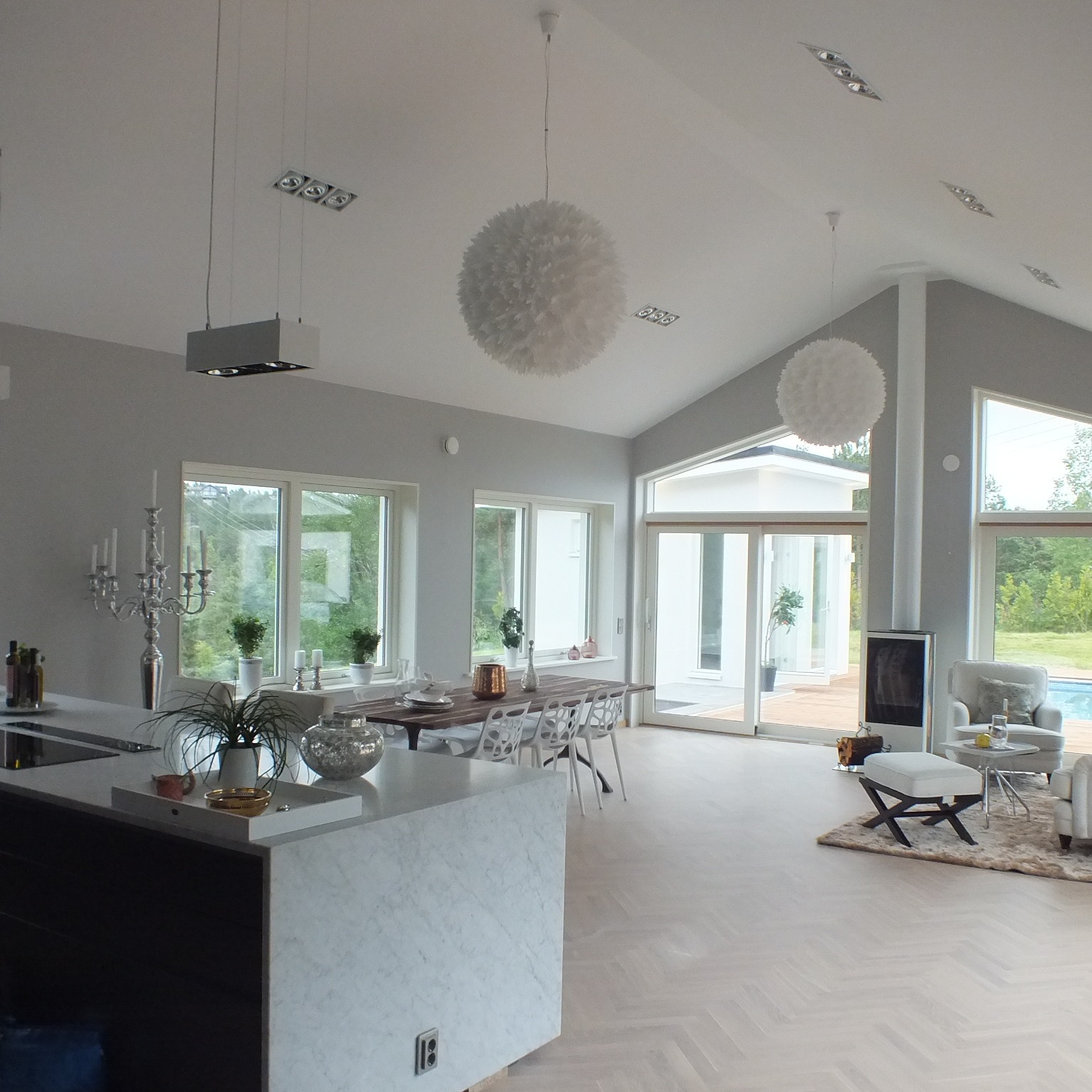 Jasne, przestronne wnętrze domu z dużymi oknami, widokiem na zewnątrz, nowoczesną kuchnią z marmurowym blatem, jadalnią i strefą relaksu z kominkiem oraz wyjściem na taras z basenem.