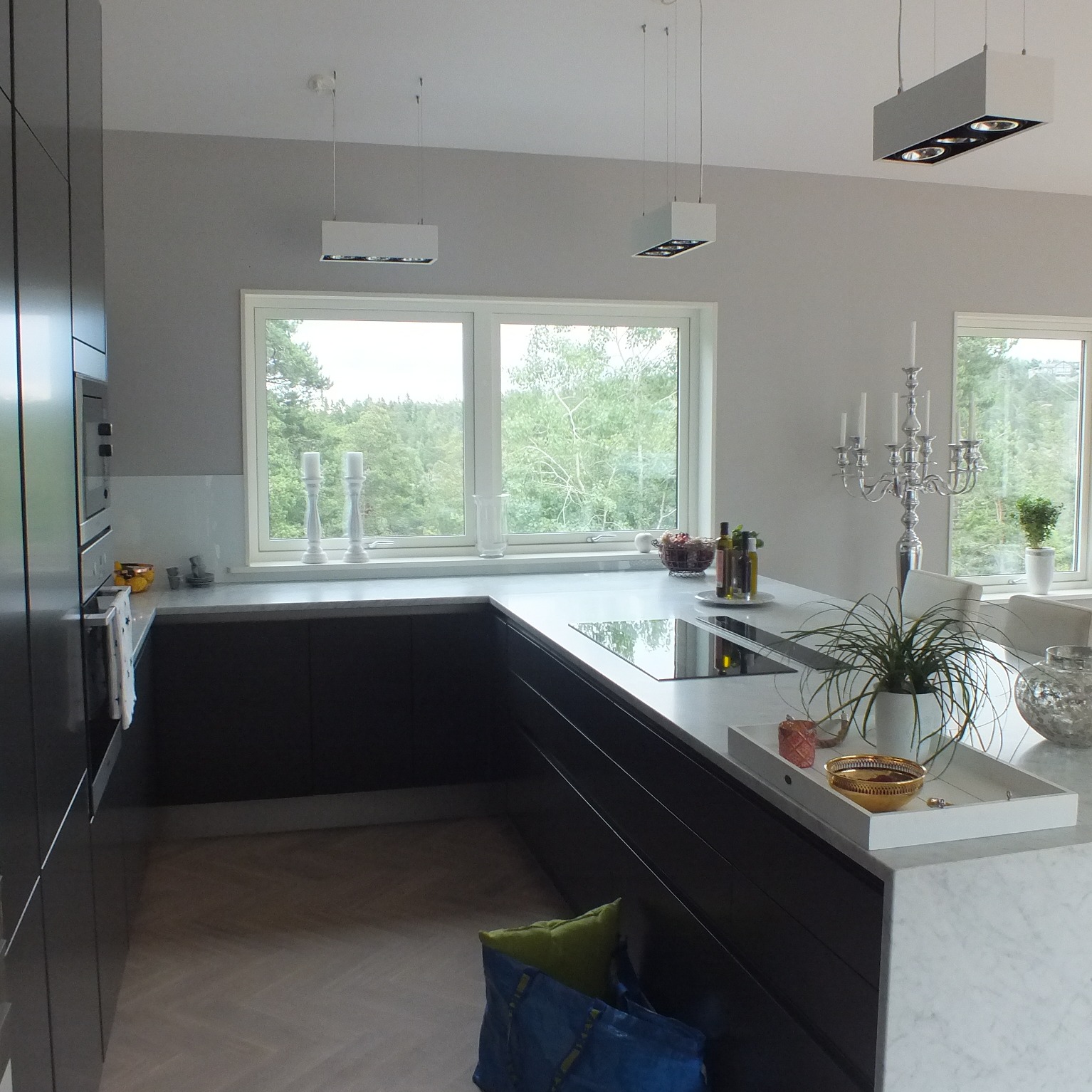 Nowoczesna kuchnia z ciemnymi szafkami i białym blatem, widok na okno z zielenią na zewnątrz, designerskie oświetlenie sufitowe i dekoracje na blacie.