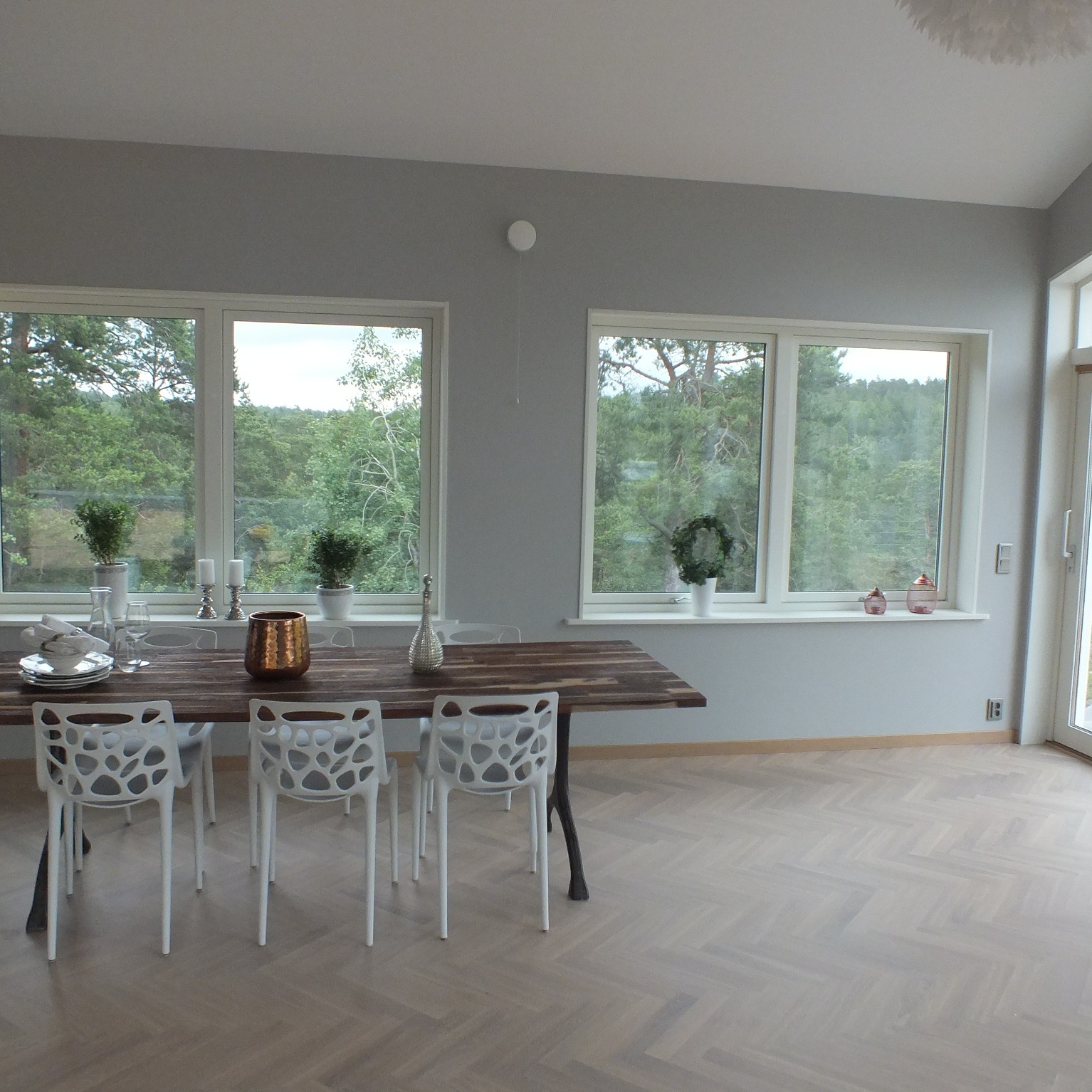 Jasne wnętrze jadalni z widokiem na las przez dwa duże okna, stół z ciemnego drewna z dekoracjami i nakryciem, białe krzesła o ażurowym wzorze.