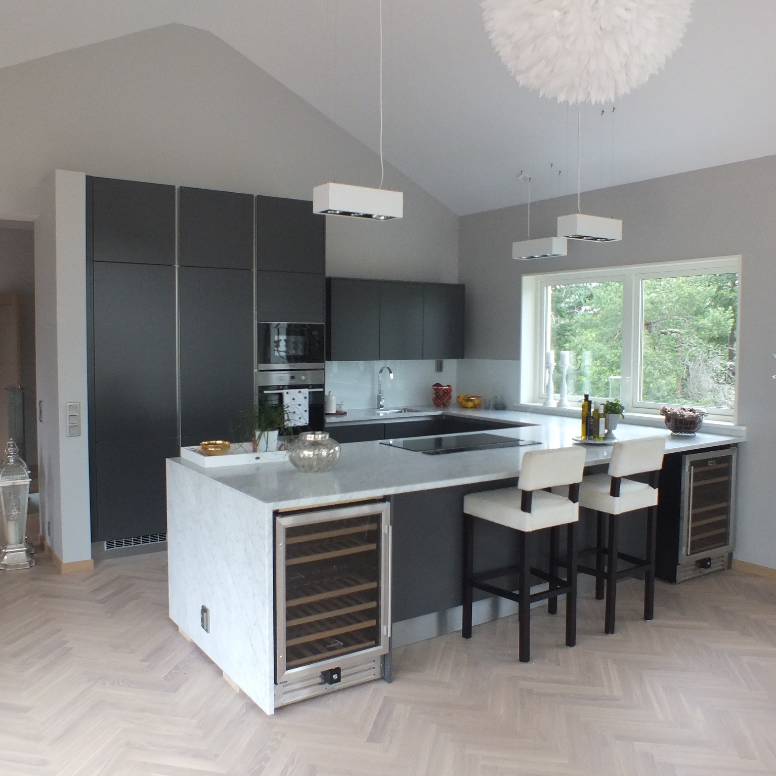 Nowoczesna kuchnia z szarymi szafkami, marmurowym blatem wyspy i wbudowaną chłodziarką do wina; podłoga z jasnego parkietu ułożonego w jodełkę.