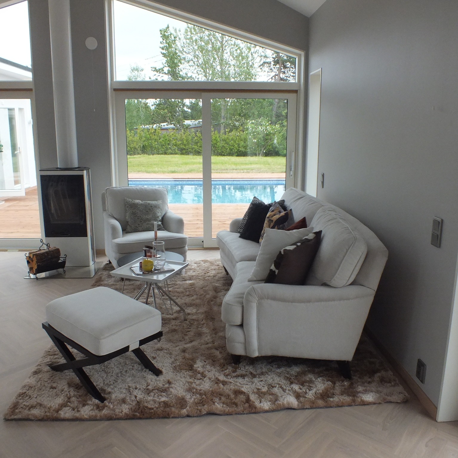Jasny salon z dużym oknem wychodzącym na basen, kominek z drewnem, białe meble, puszysty dywan i parkiet ułożony w jodełkę.