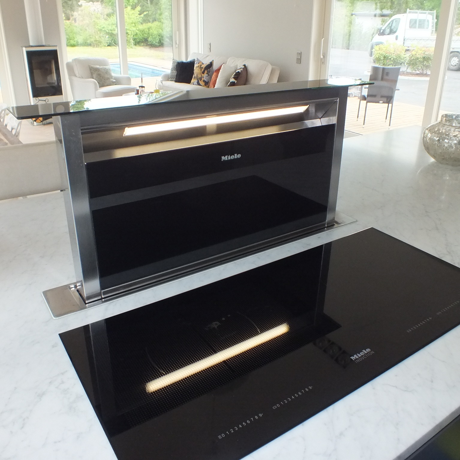 Nowoczesna płyta indukcyjna Miele z wysuwanym okapem, umieszczona na blacie z białego marmuru, z widokiem na salon i basen przez duże okno.