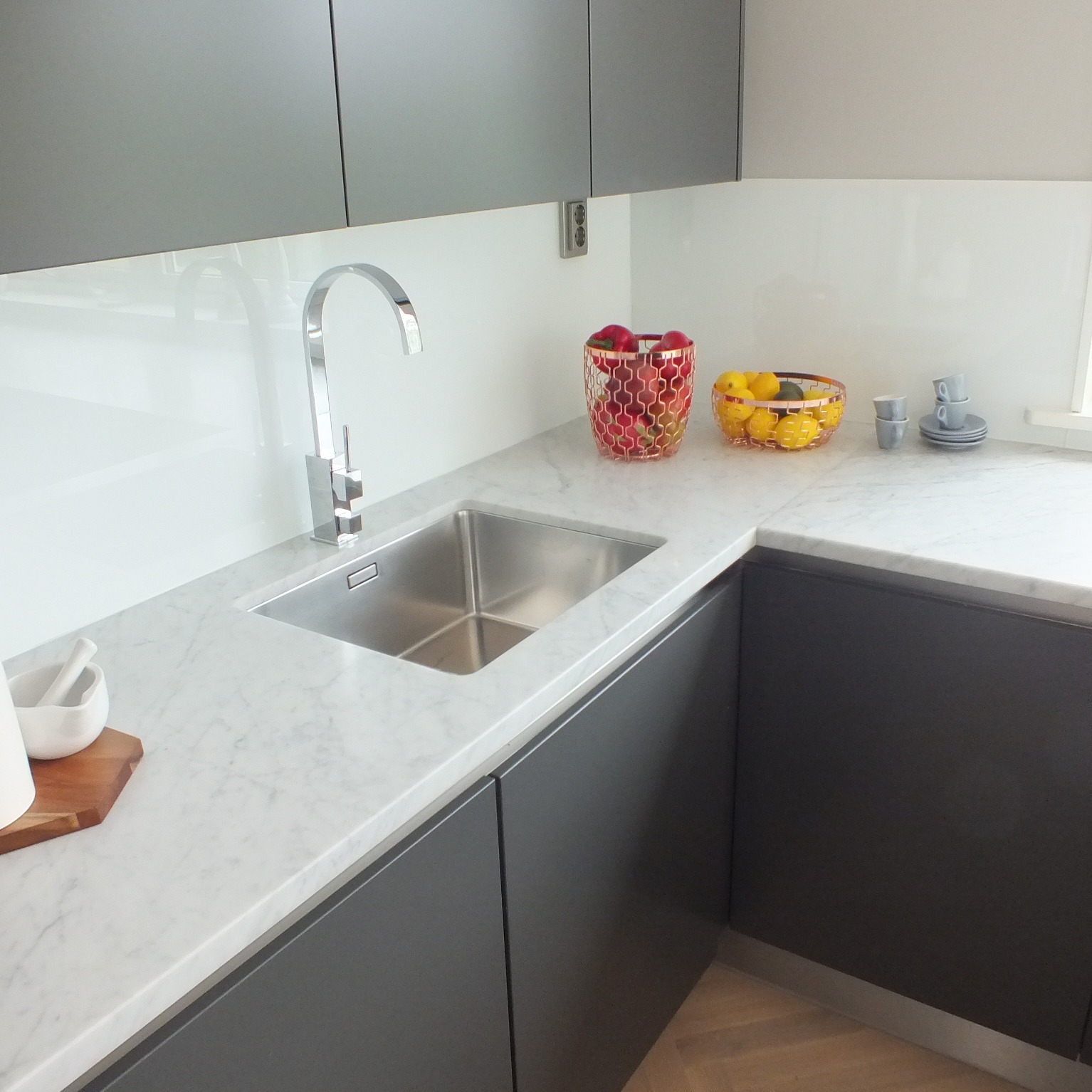 Nowoczesna kuchnia z szarymi szafkami, marmurowym blatem i stalowym zlewozmywakiem. Na blacie koszyki z owocami i filiżanki.