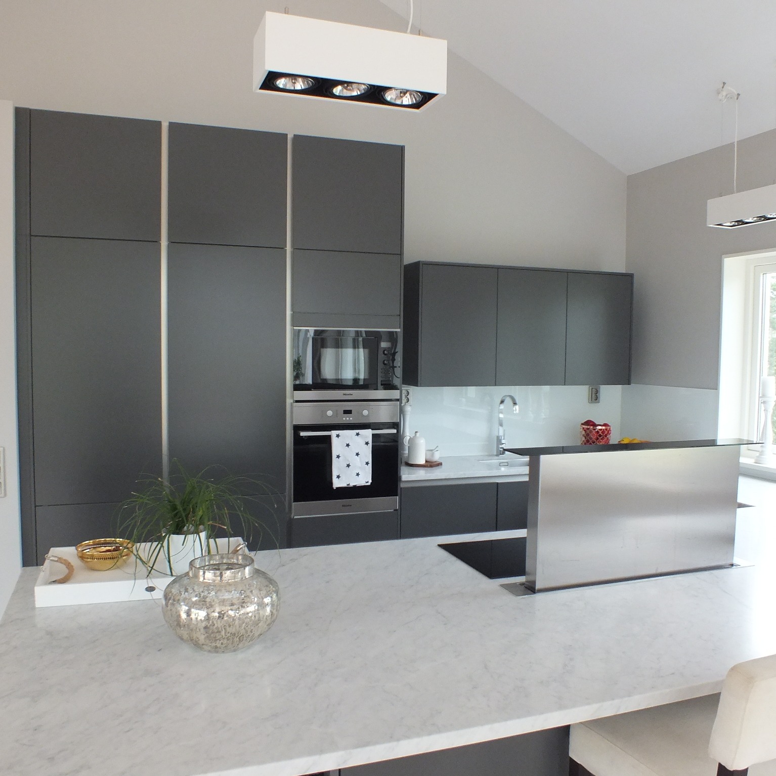 Nowoczesna kuchnia z szarymi szafkami, marmurowym blatem i srebrną wysuwaną okapem, oświetlona białymi lampami sufitowymi.