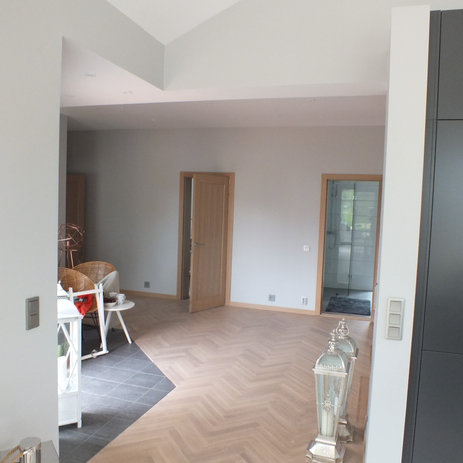 Jasne wnętrze z parkietem w jodełkę, dwoje drzwi z drewnianymi ościeżnicami, widok na łazienkę z kabiną prysznicową oraz fragment aranżacji salonu z wiklinowymi fotelami.