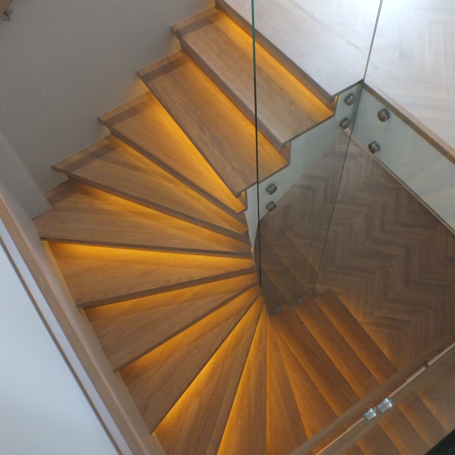 Kątowe ujęcie drewnianych schodów spiralnych z podświetleniem LED i szklaną balustradą mocowaną za pomocą metalowych śrub.