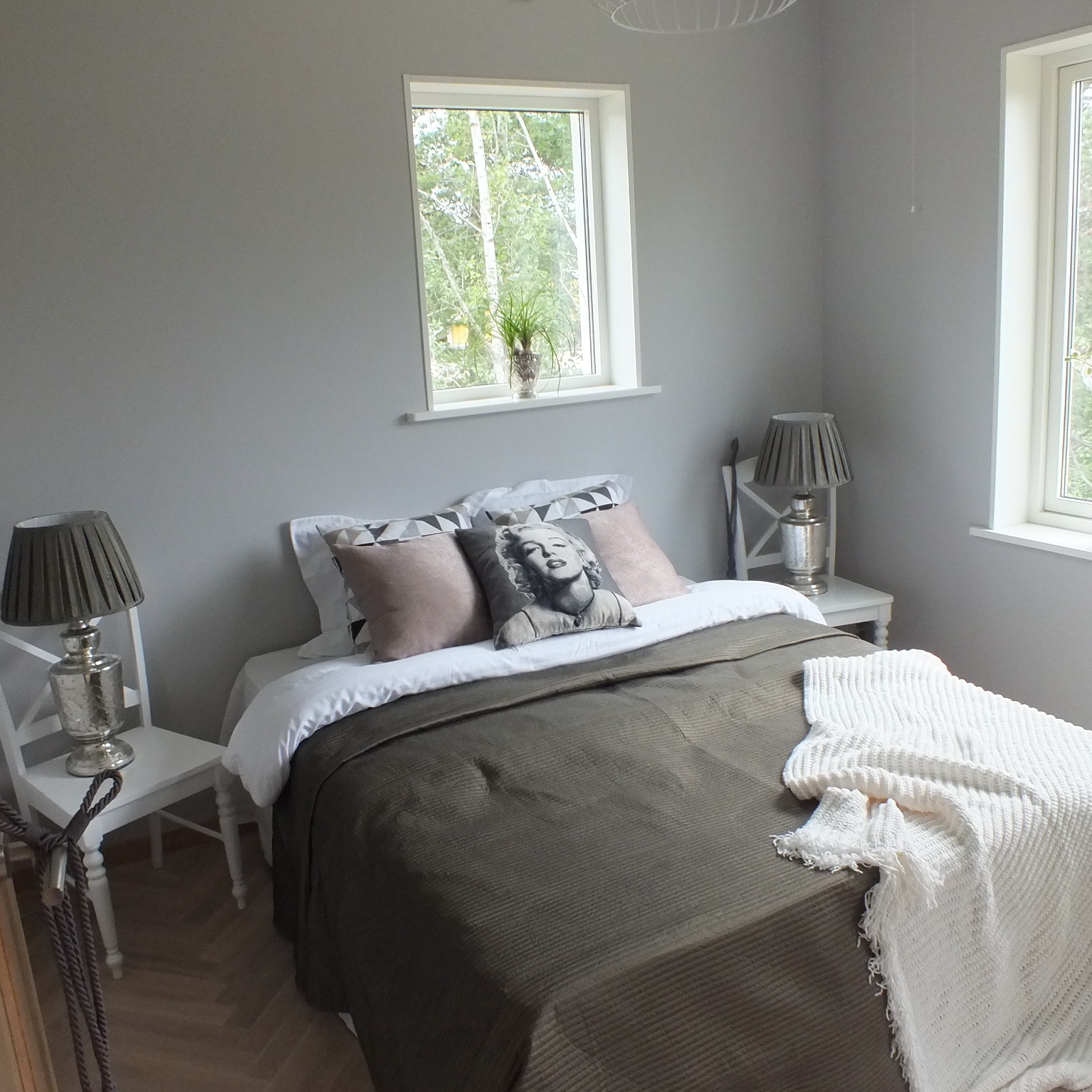 Jasnoszara sypialnia z łóżkiem z brązową narzutą, białym pledem i poduszką z Marilyn Monroe, białe okna z widokiem na zieleń, lampki nocne na białych krzesłach