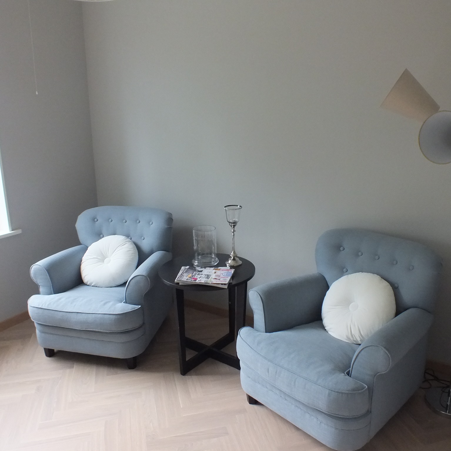 Dwa jasnoniebieskie fotele z okrągłymi białymi poduszkami na siedziskach, ustawione symetrycznie po obu stronach małego, okrągłego, czarnego stolika z czasopismami i szklanymi naczyniami,...