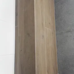 Ułożone pionowo płytki imitujące drewno w odcieniach brązu sąsiadują z białymi płytkami po lewej i szarymi płytkami o teksturze betonu po prawej stronie.