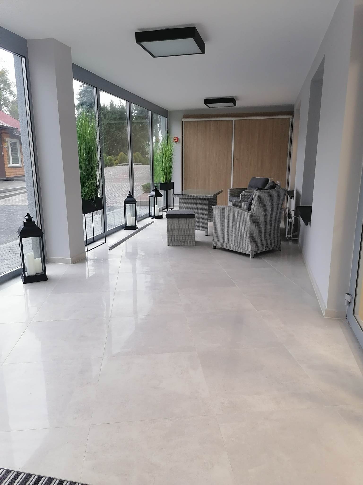 Jasne, błyszczące płytki podłogowe w przeszklonym pomieszczeniu z meblami ogrodowymi z rattanu, dekoracyjnymi lampionami i wysokimi trawami w donicach.