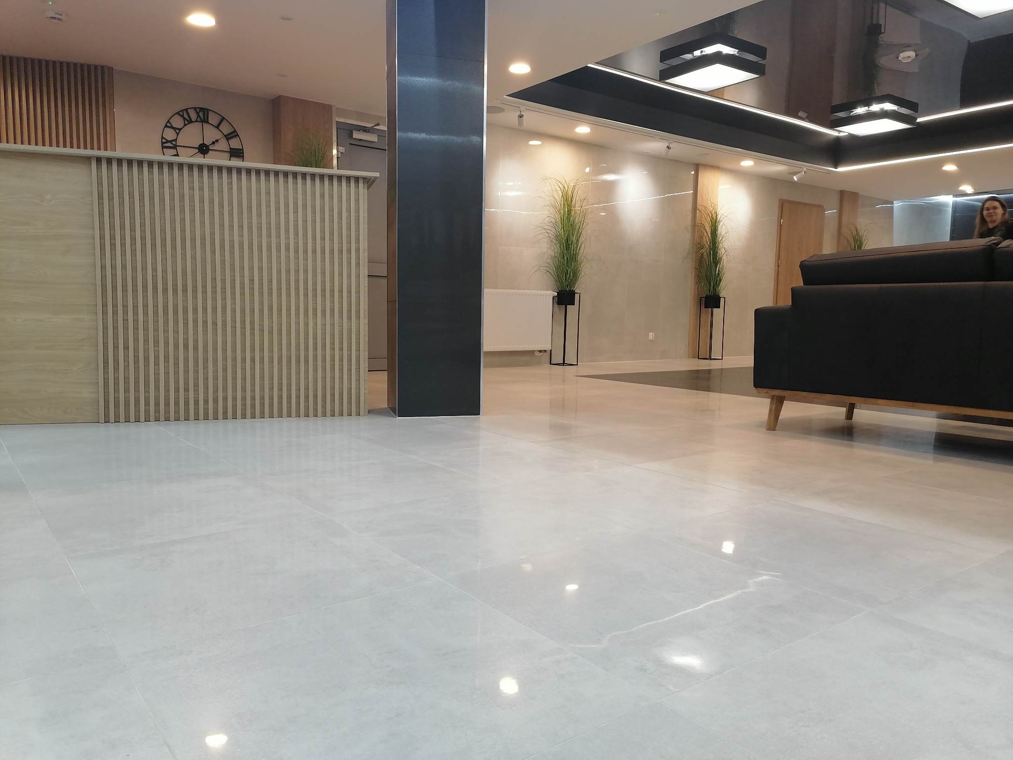 Błyszcząca, jasnoszara podłoga z płytek ceramicznych w nowoczesnym holu z elementami drewna i czerni; widoczne rośliny w wysokich donicach, recepcja z zegarem i część sofy.