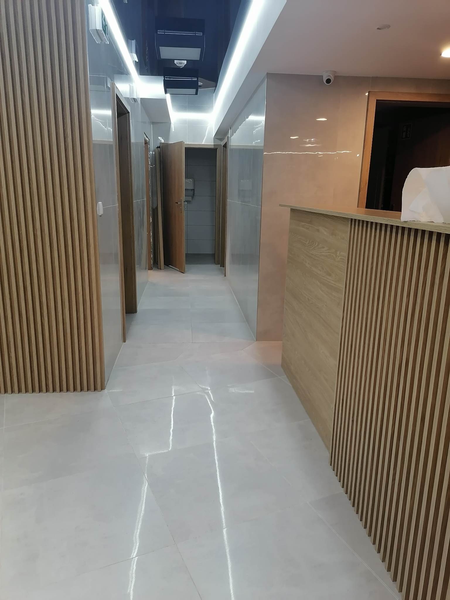 Korytarz wykończony płytkami w odcieniach beżu i szarości z drewnianymi panelami dekoracyjnymi, oświetleniem LED i otwartymi drzwiami do pomieszczeń.