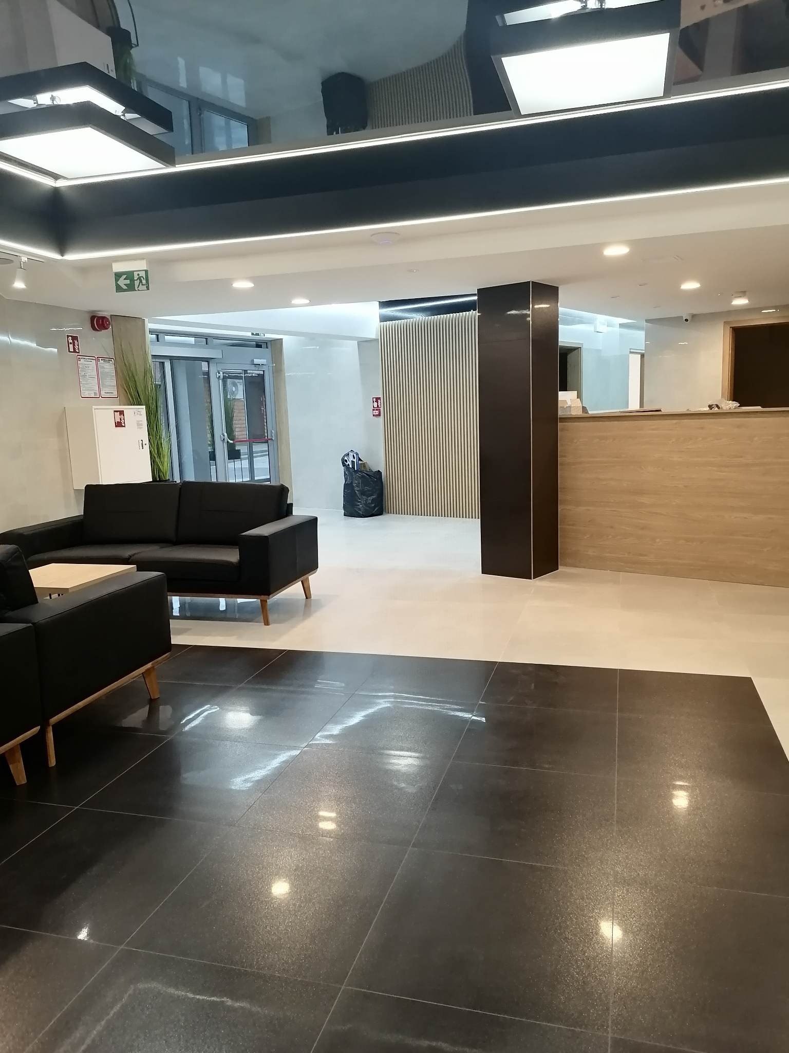Nowoczesne wnętrze holu z błyszczącą, ciemną podłogą z dużych płytek, czarnymi sofami i recepcją z drewnianym wykończeniem.